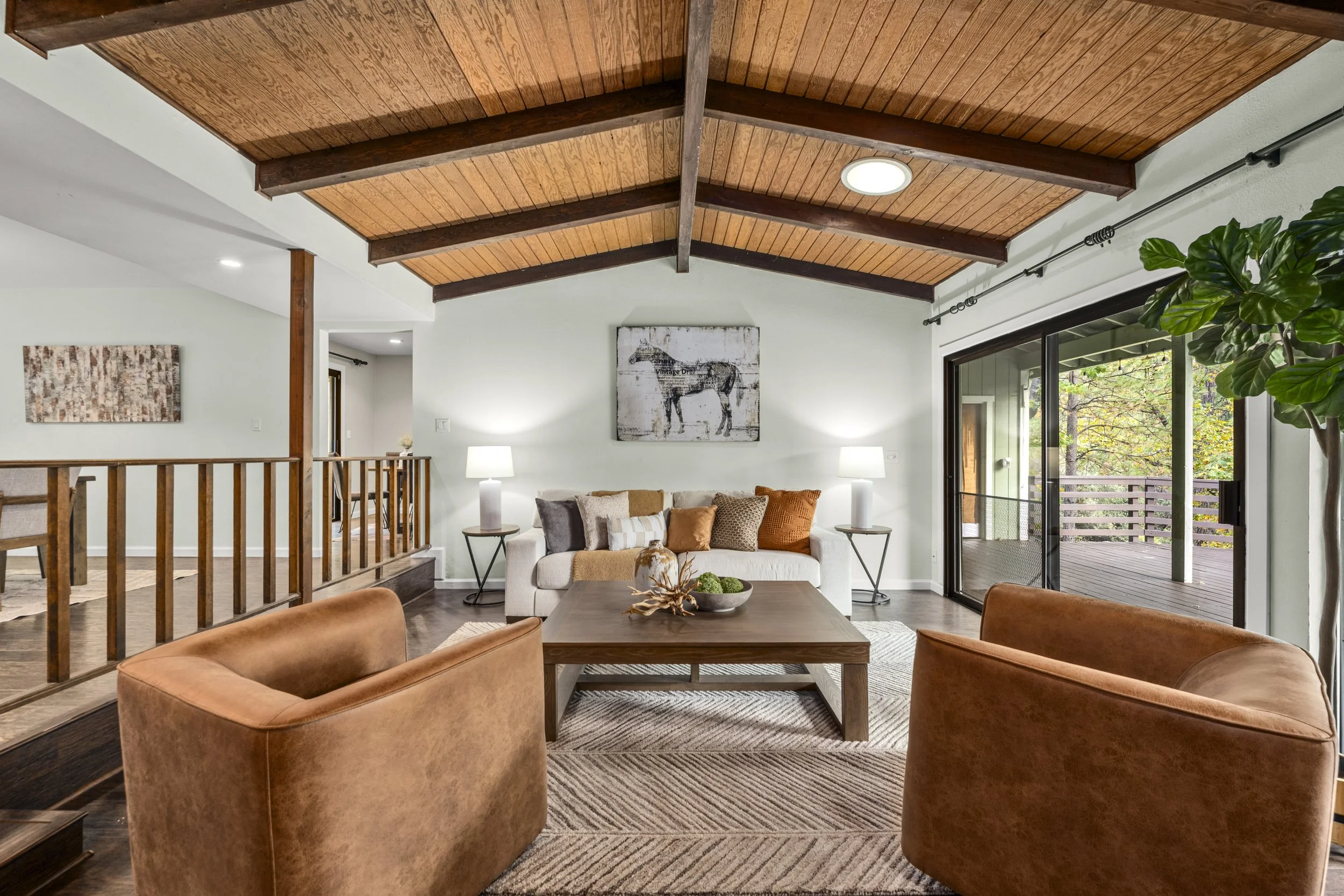 Living room with a wooden vaulted ceiling, white walls, a beige sofa with orange and beige pillows, a wooden coffee table with a decorative item, tan armchairs, large sliding glass door leading to outdoor deck, and artwork on the walls.