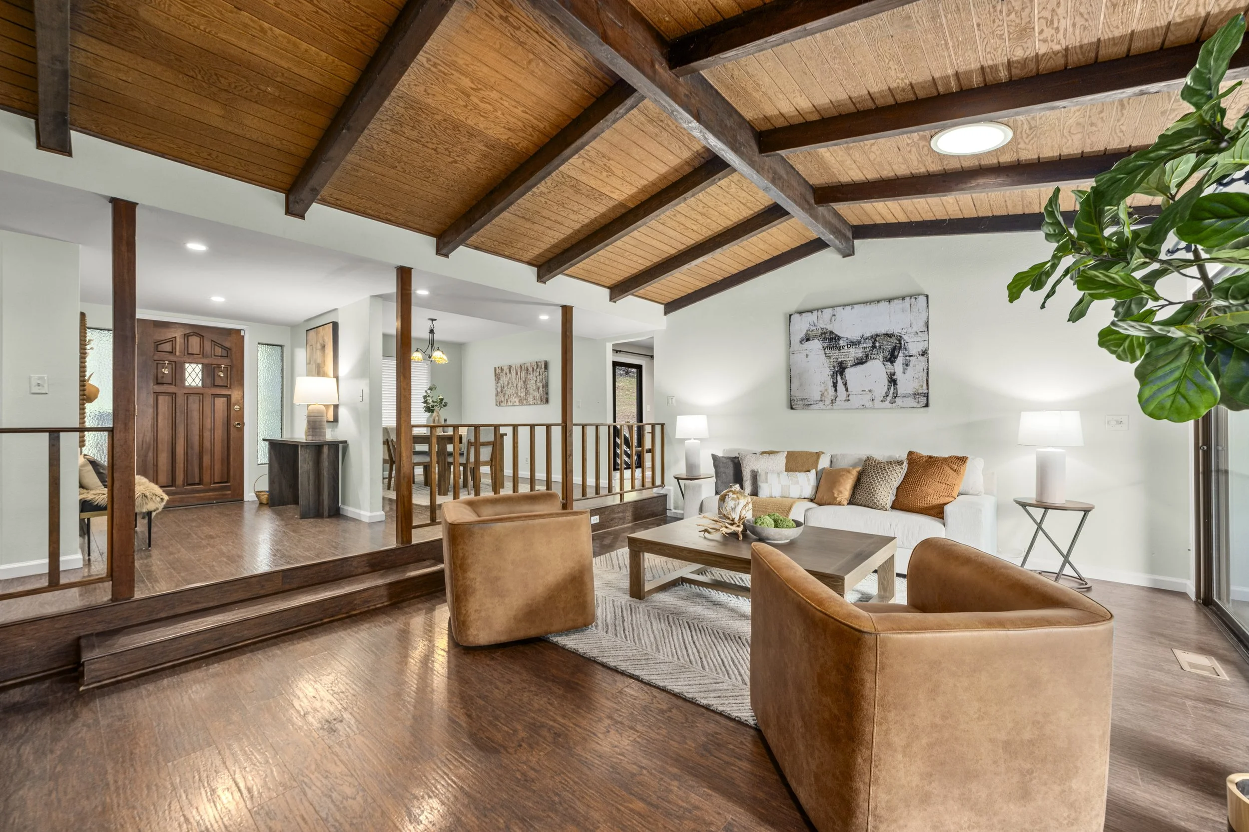 Living room with brown leather armchairs, wooden coffee table, white sofa with pillows, and a large plant. Wooden ceilings with beams and recessed lighting, hardwood flooring, and wall art. Entry door and dining area visible in the background.