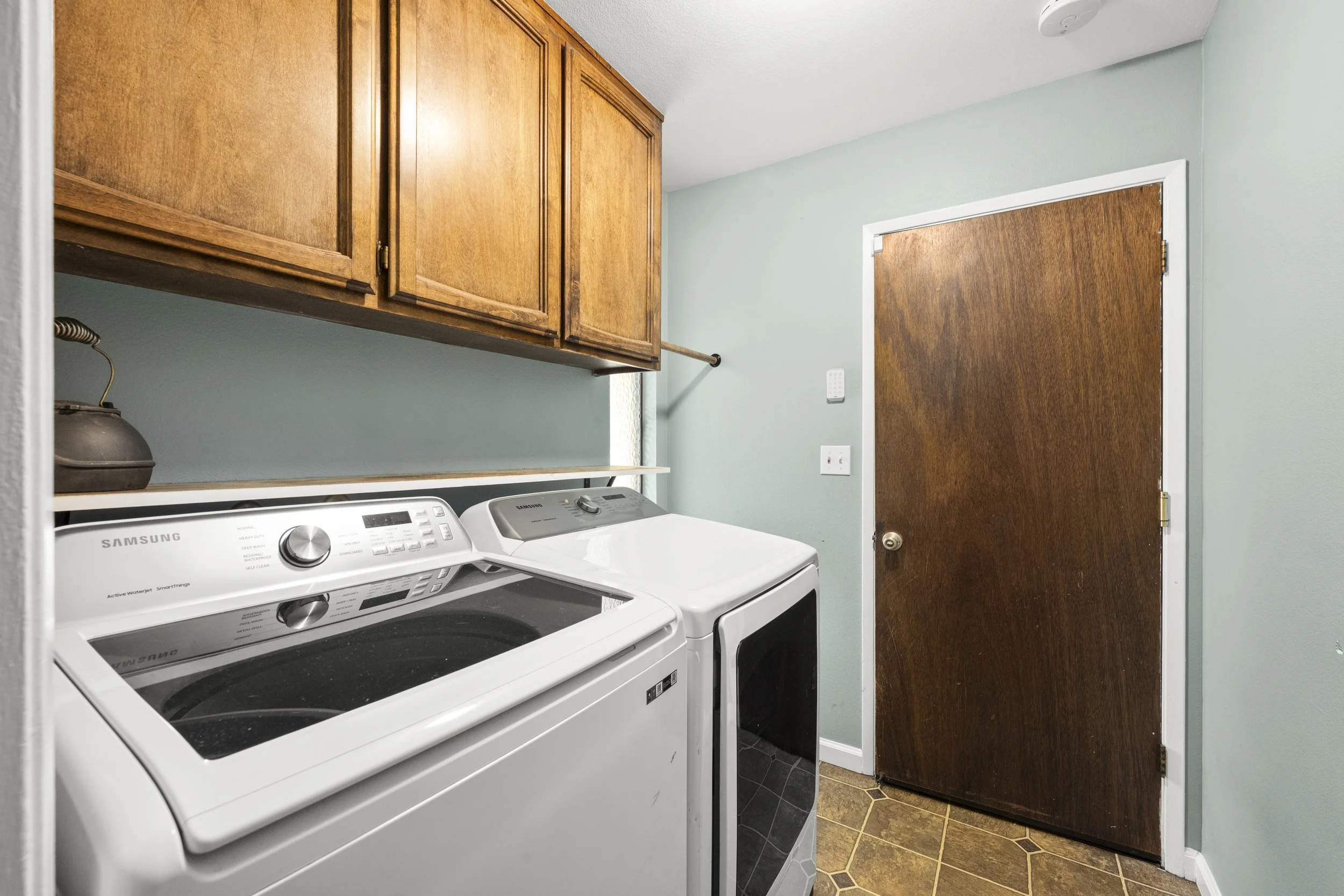A laundry room with two white washing machines, wooden cabinets, a brown door, and a section of tiled flooring.