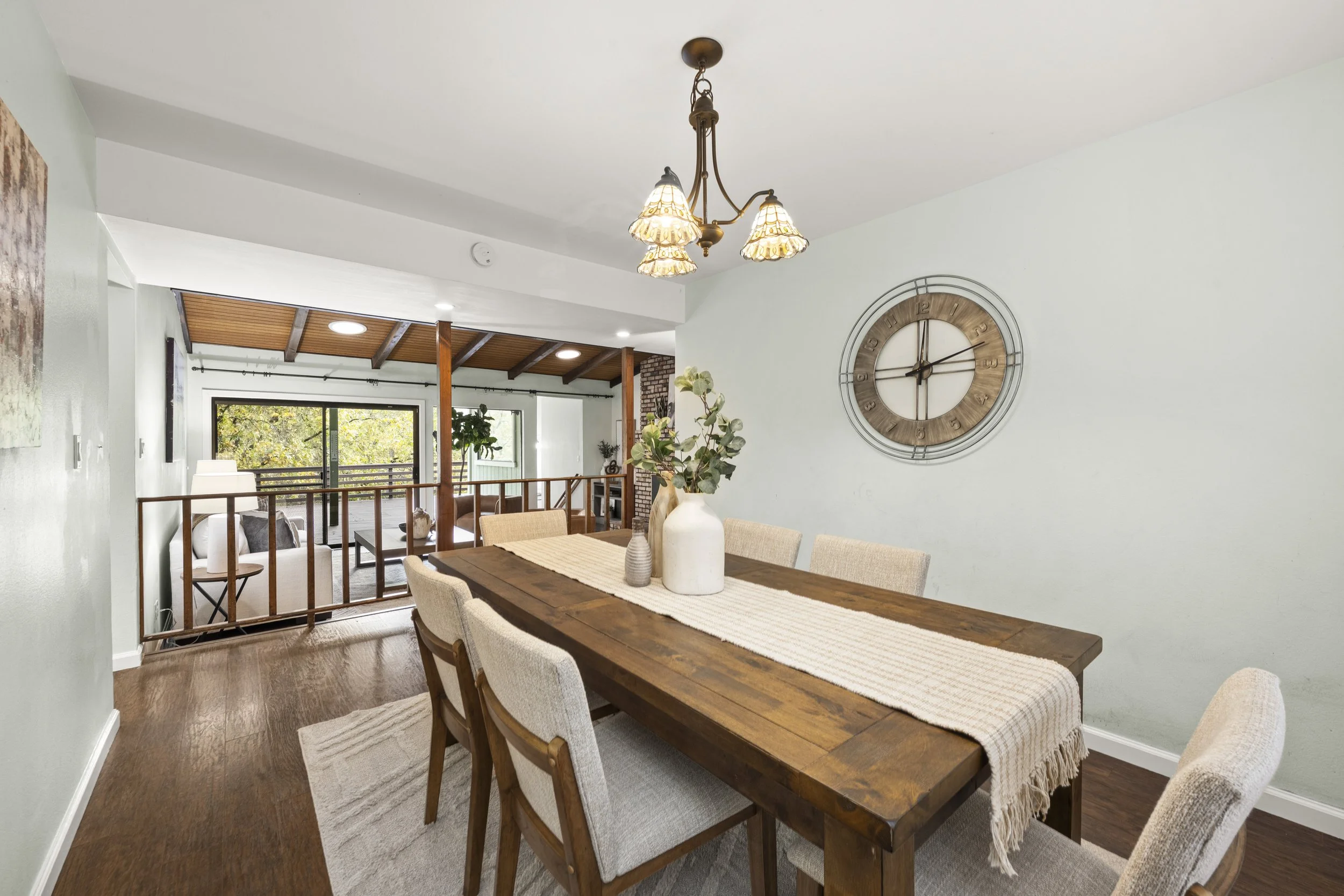 Dining room with a large wooden table, beige upholstered chairs, and vases with greenery. A wall clock and chandelier ornaments are visible. The room features wood flooring and overlooks a living space with large windows.