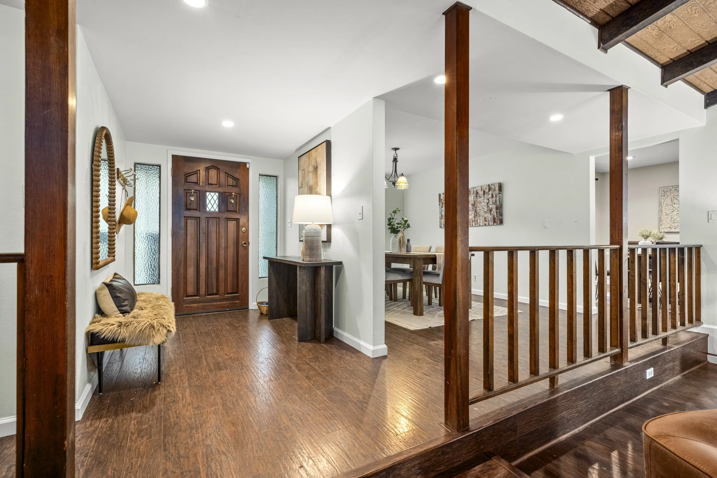 Living room entryway with wooden front door, glass side windows, dark hardwood flooring, a small fur-covered bench with pillows, a side table with a lamp, and a dining area with a wooden table and chandelier in the background.