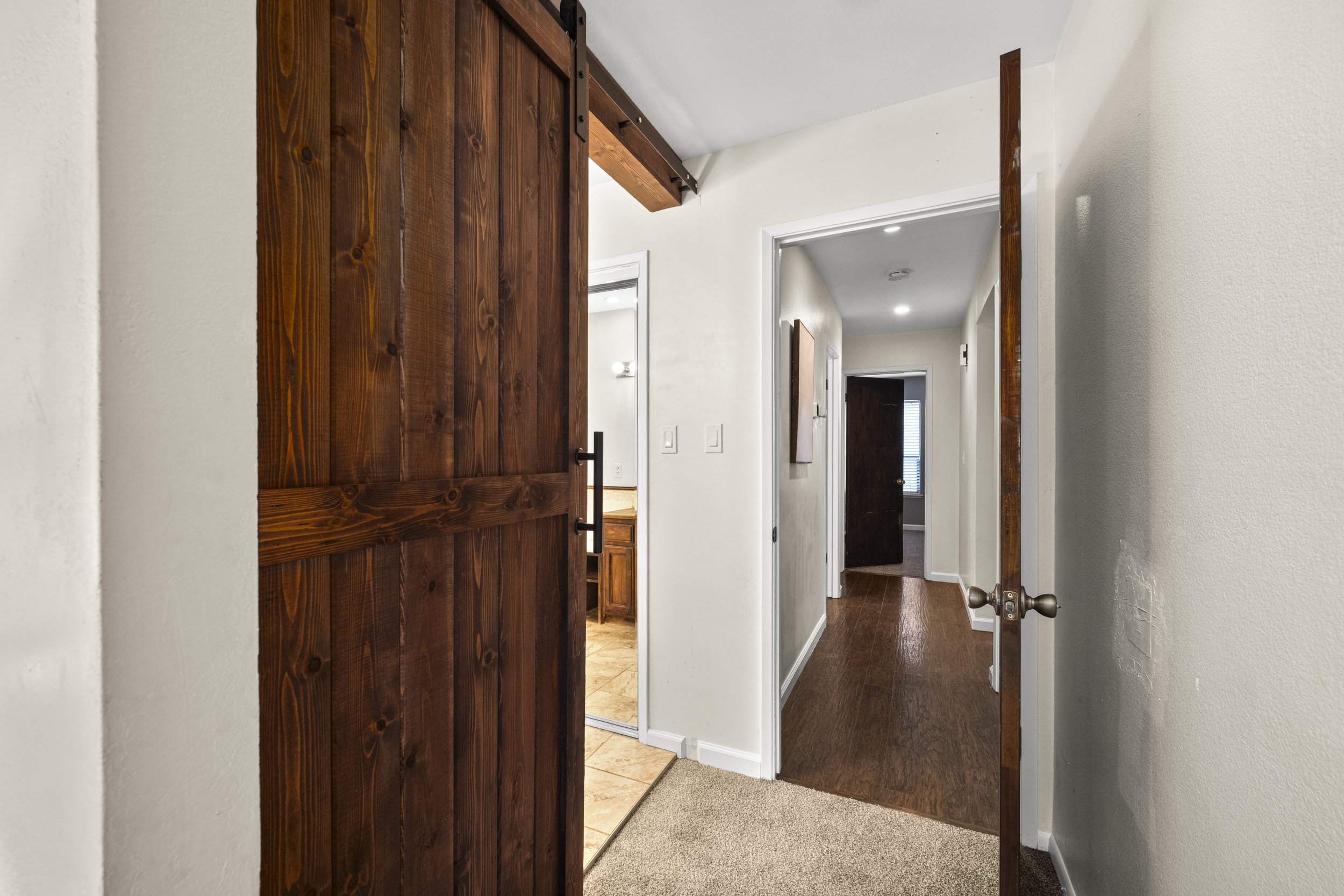 Open wooden sliding door reveals a hallway with hardwood floors and white walls, with doors on either side and a closet or room visible in the background.