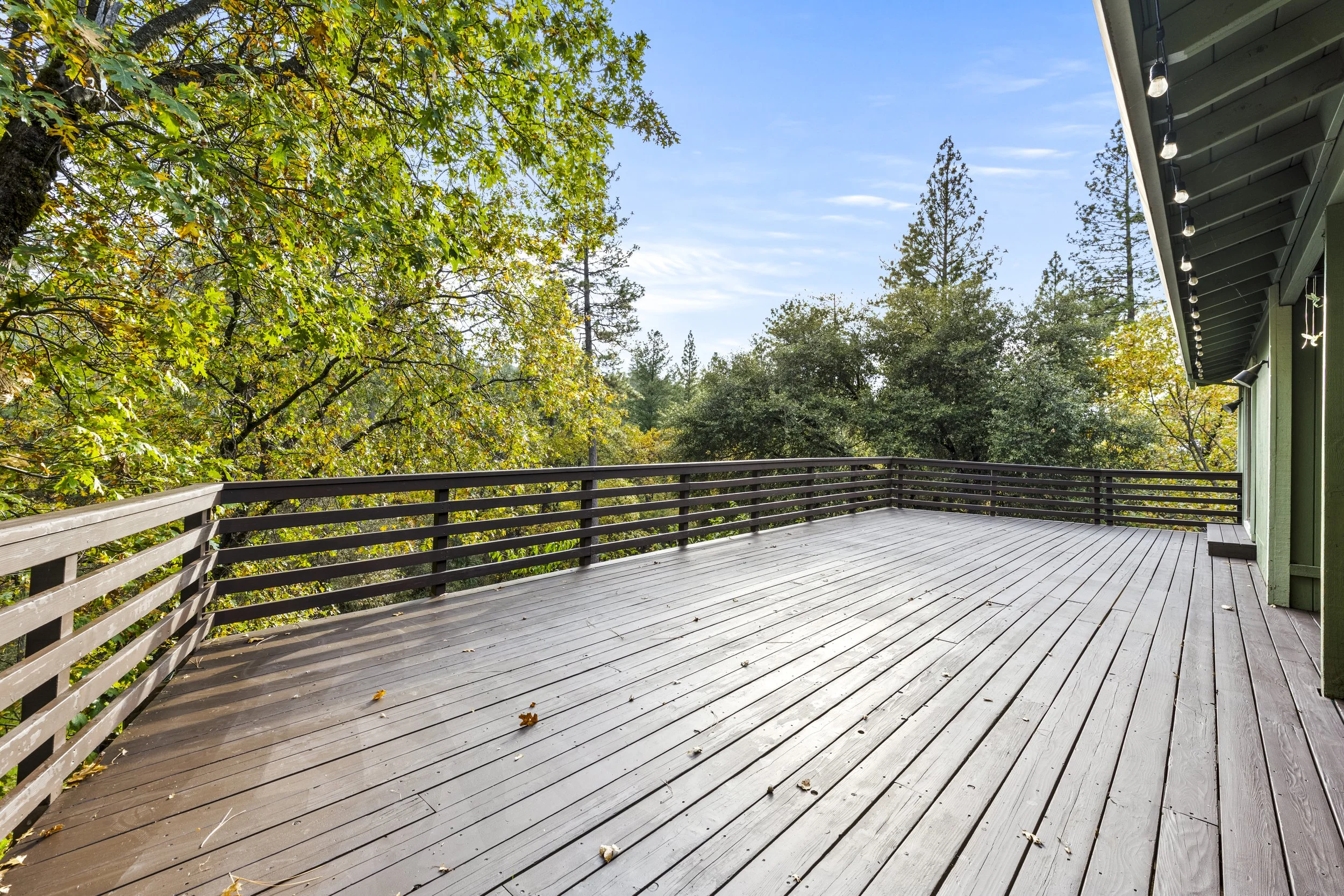 View of a spacious outdoor wooden deck attached to a house with a green exterior, overlooking lush trees with fall foliage and a clear blue sky.