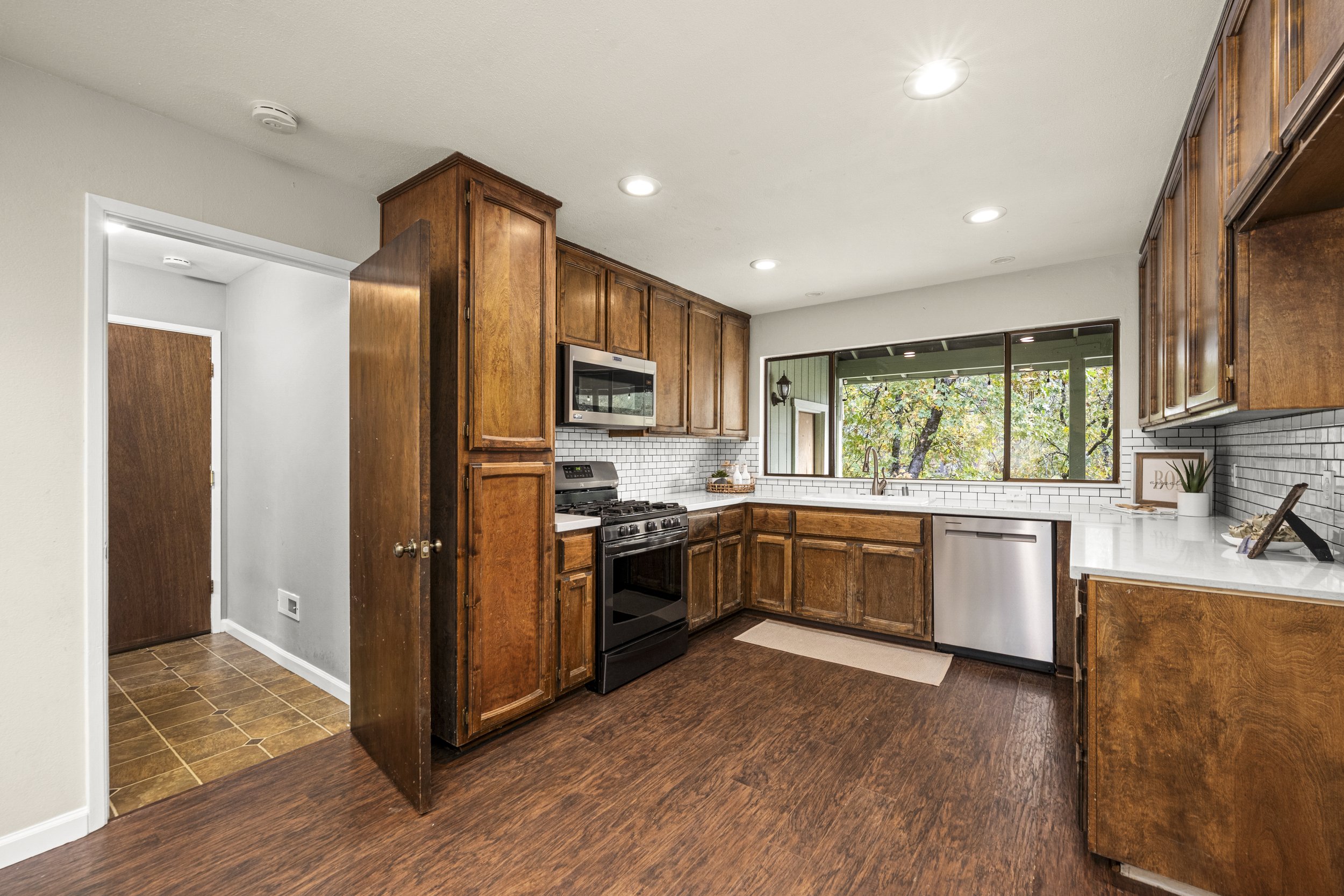 A kitchen featuring wooden cabinets, a white countertop, a stainless steel oven and microwave, a window overlooking trees, and a white subway tile backsplash.