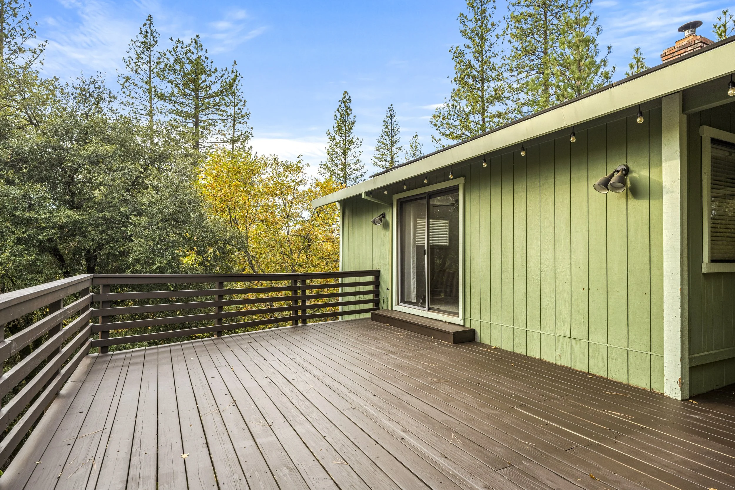 A spacious wooden balcony attached to a house with green siding, surrounded by tall pine trees and other foliage, under a blue sky with some clouds.