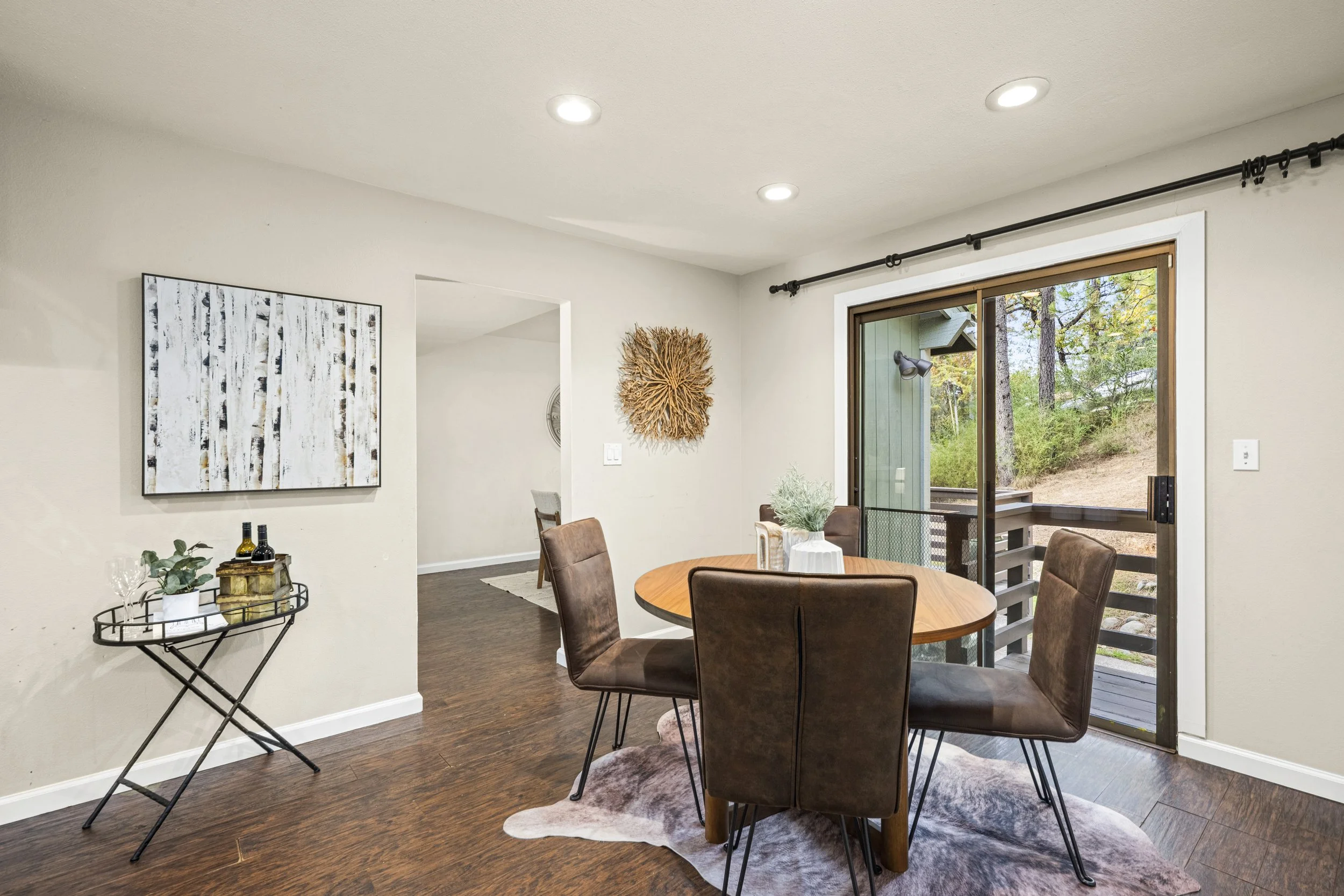 A dining area with a round wooden table and six brown chairs, a sliding glass door showing an outdoor view of trees, a wall decorated with wall art and a branch sculpture, and a small side table with wine bottles and plants.