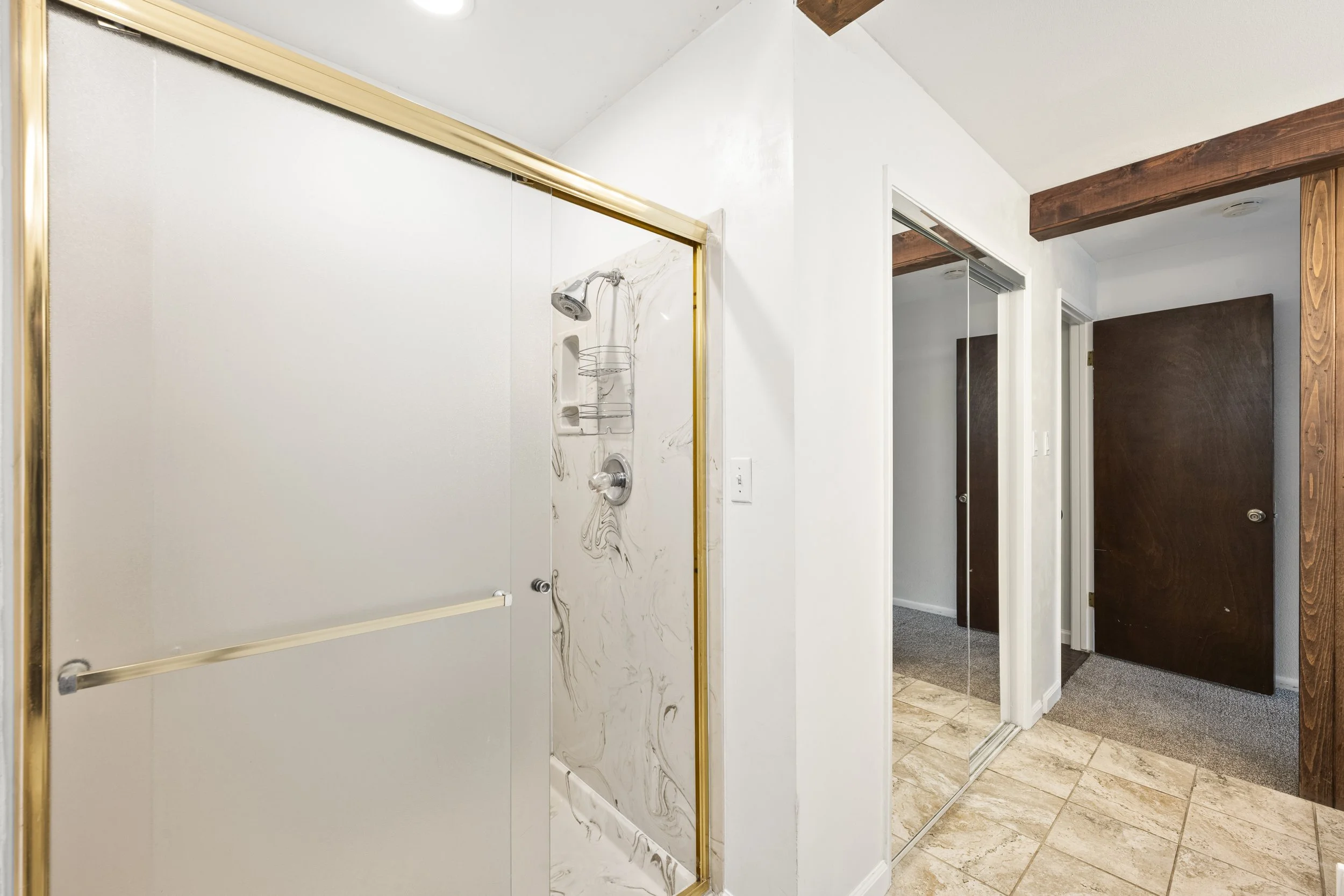 Bathroom with a shower stall on the left, featuring a marble wall, a showerhead, and a glass door with gold trim. To the right, there's a large mirror wall reflecting part of the room, with dark brown wooden beams above. The room has beige tiled floo