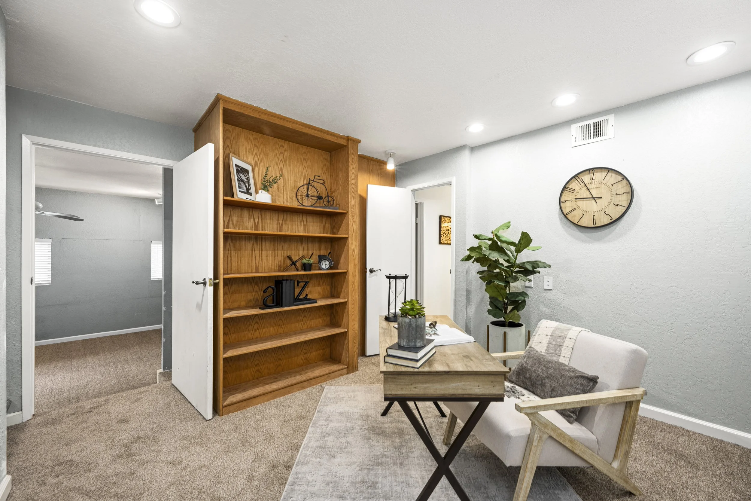 Living room with a wooden bookshelf, a wall clock, a potted plant, and a cozy seating area with cushions.