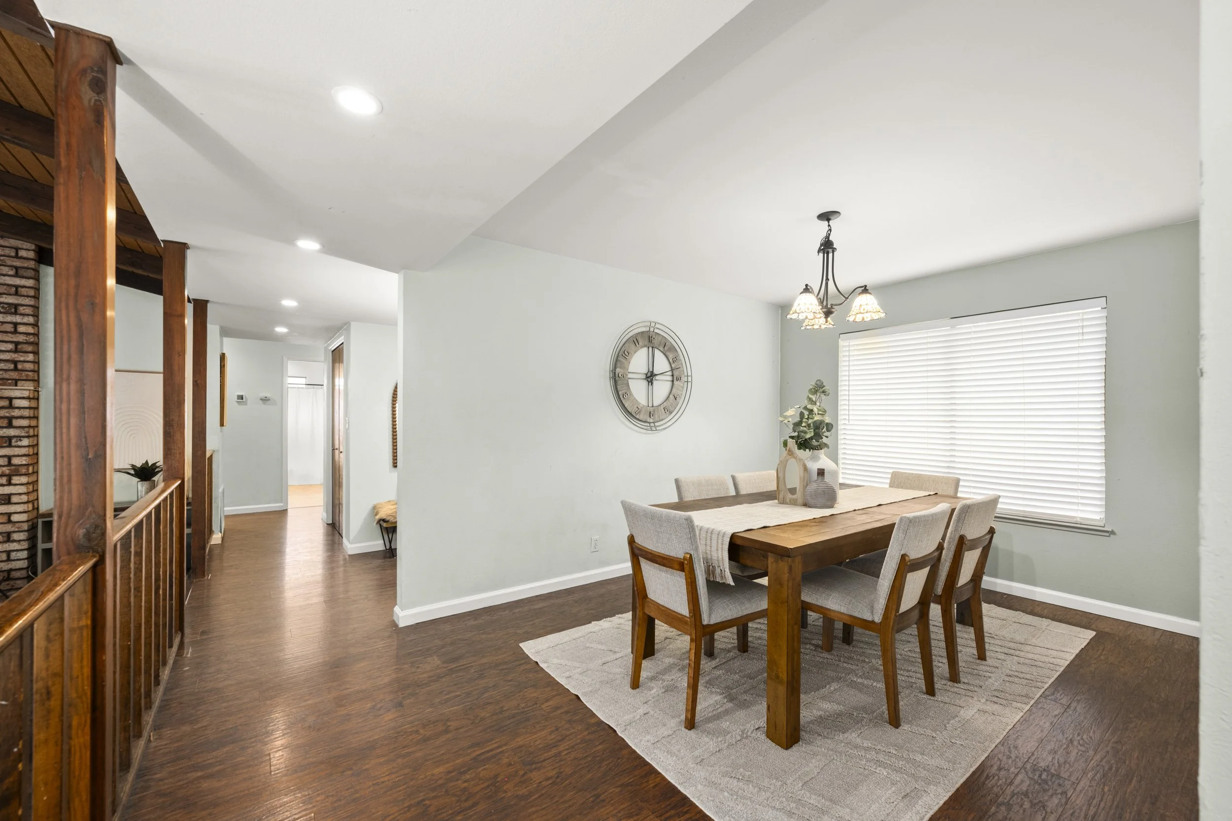 Living and dining room with wooden dining table and six upholstered chairs, wall clock, window with blinds, overhead chandelier, and hardwood flooring.