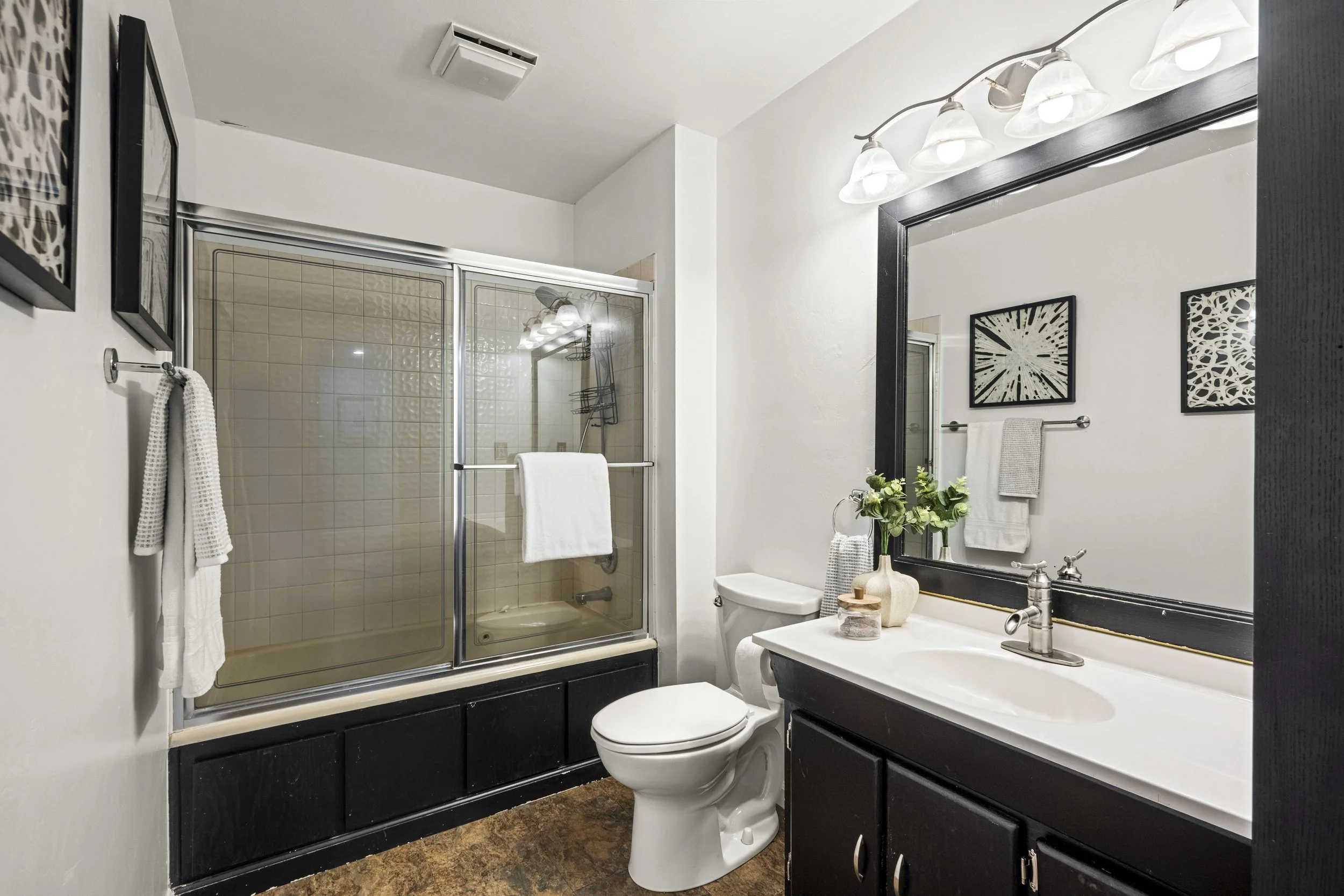 A modern bathroom with a glass-enclosed shower, a white toilet, a white sink with a black cabinet, a large mirror with a black frame, and wall decor. There are towels hanging and a small plant on the counter. Lighting is provided by four overhead lig