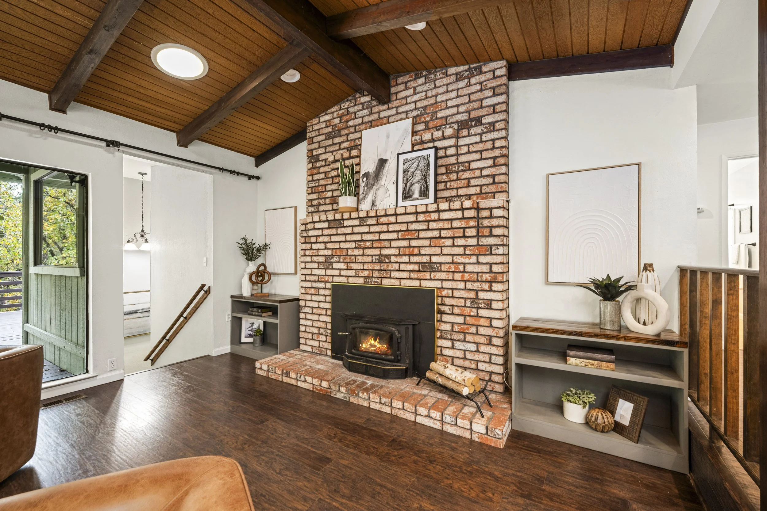 Living room with brick fireplace, wood ceiling beams, and art decor. Open door shows trees outside.