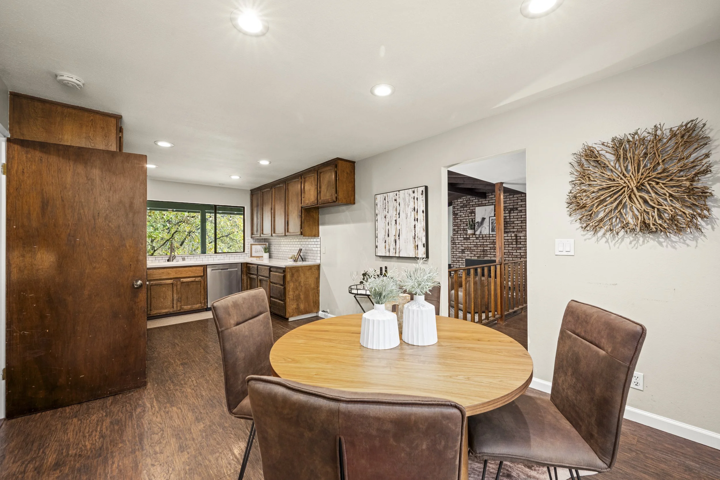View of a kitchen and dining area with round wooden table and four brown upholstered chairs, decorative vases on table, wooden cabinetry, window with greenery outside, wall art, driftwood wall sculpture, and brick accent wall.