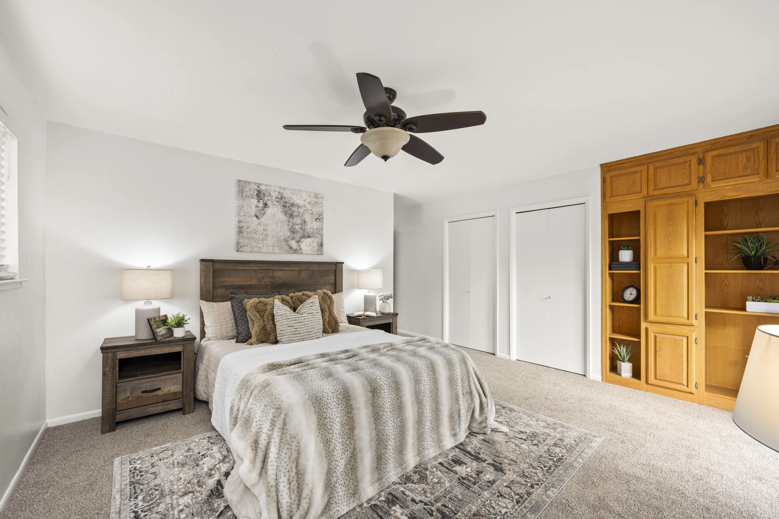 A cozy bedroom with a wooden bed, a nightstand with lamps and decor, a patterned area rug, a wall art, a ceiling fan, and a wooden wardrobe with plants inside.