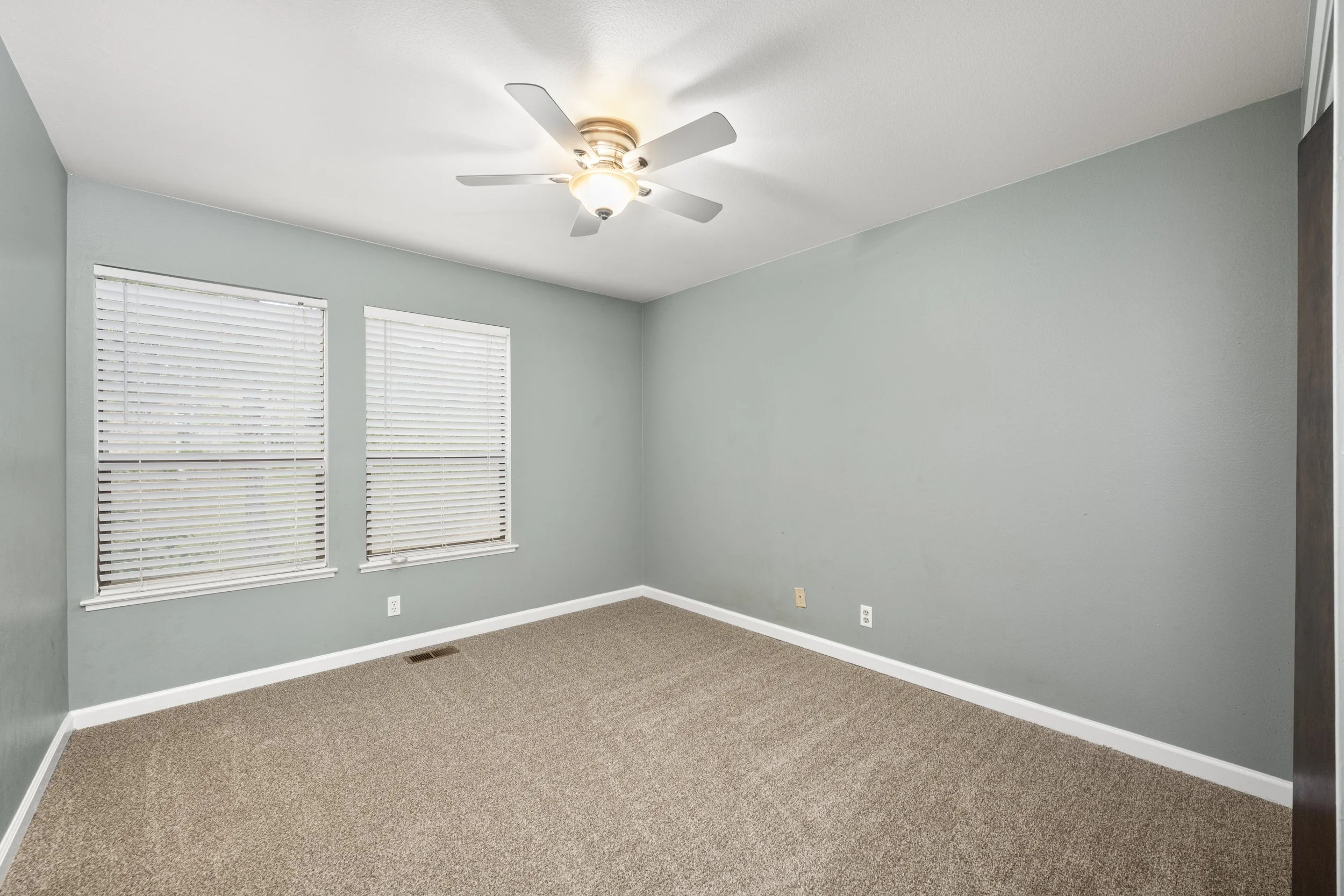 Empty room with light green walls, two windows with blinds, beige carpet, ceiling fan, and electrical outlets.