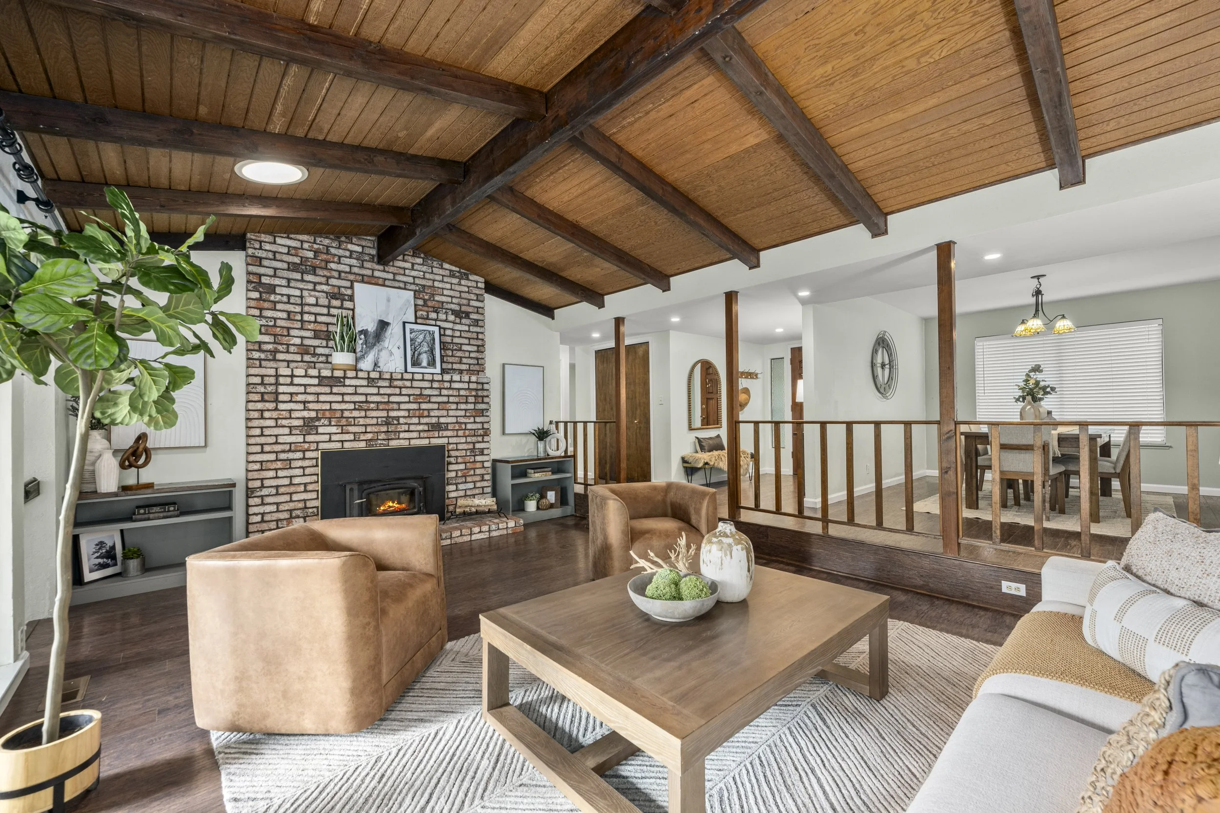 Living room with a brick fireplace, wooden ceiling, and beige furniture, including a sofa, chairs, and a coffee table with decorative items, in a cozy home interior.