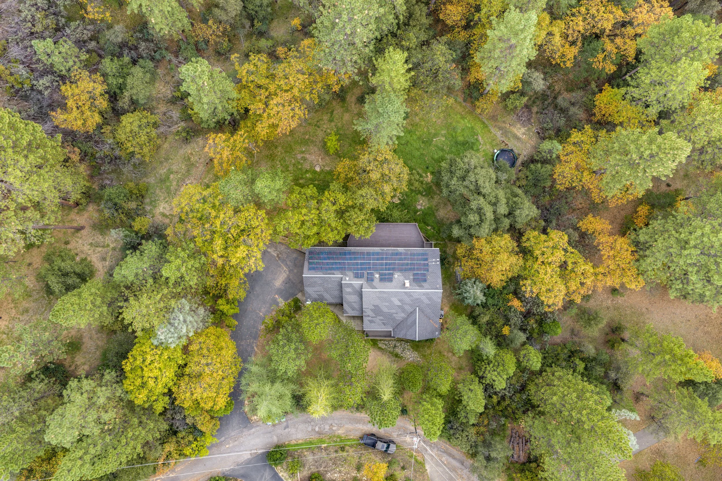 An aerial view of a house surrounded by trees with fall foliage, a lawn, a driveway with parked cars, and power lines.