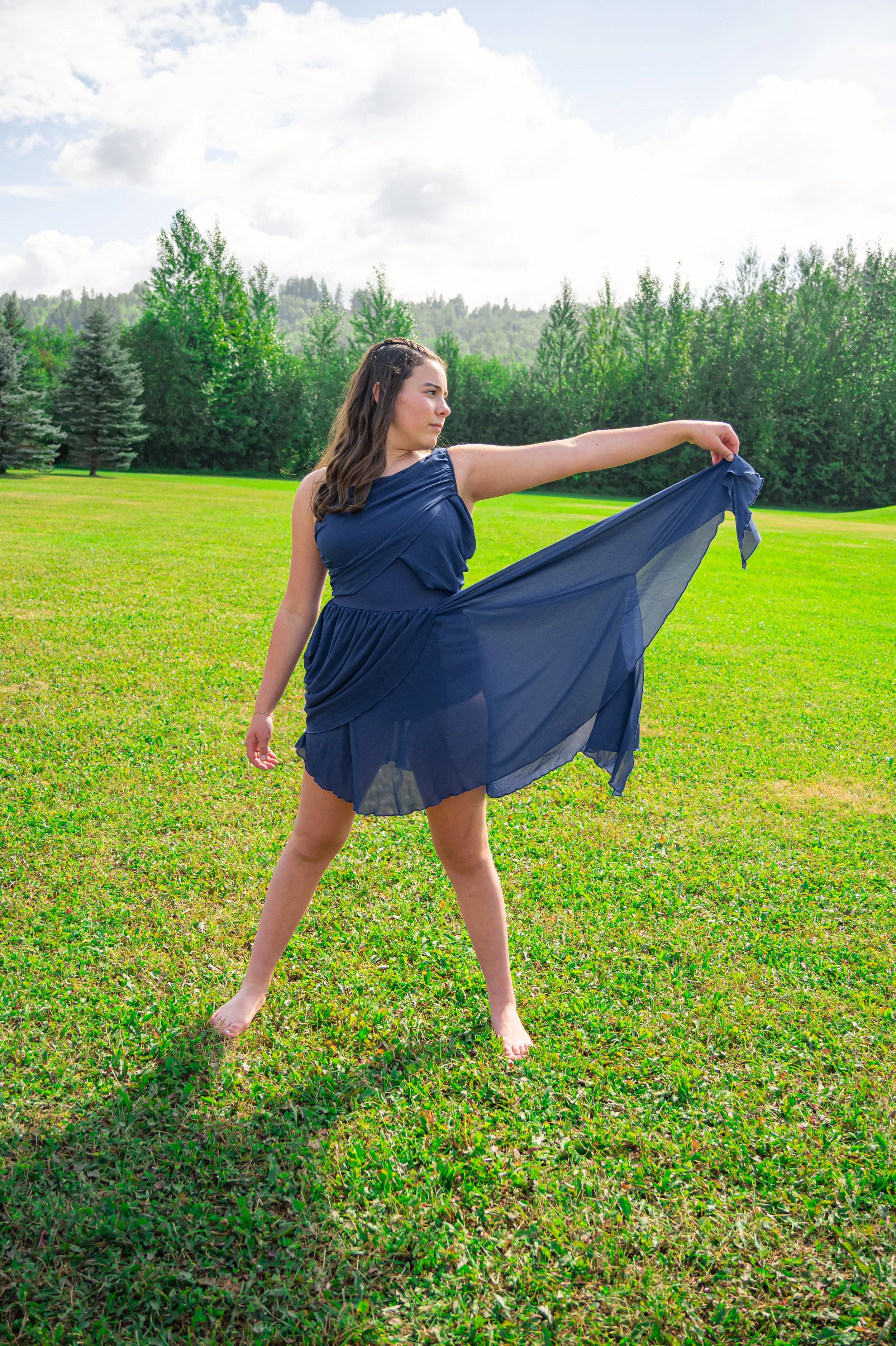 A young woman in a navy blue dress is standing barefoot on a grassy field with a backdrop of green trees and a partly cloudy sky. She is holding out her dress with her right hand and looking to her left.