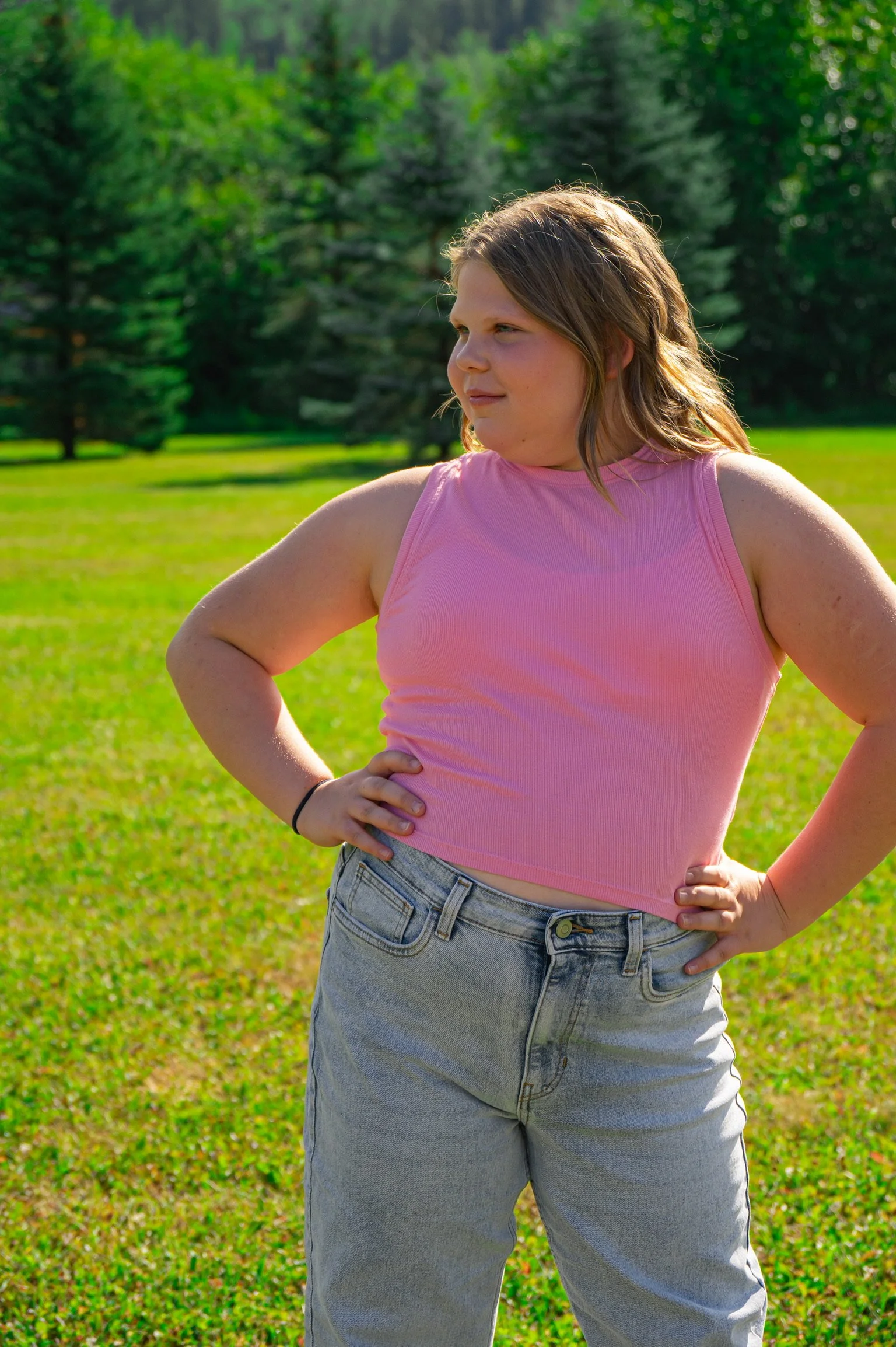 A young woman with light skin, wearing a pink sleeveless crop top and light blue jeans, stands outdoors in a grassy area with pine trees in the background. She has her hands on her hips and is looking slightly to the right with a confident expression.