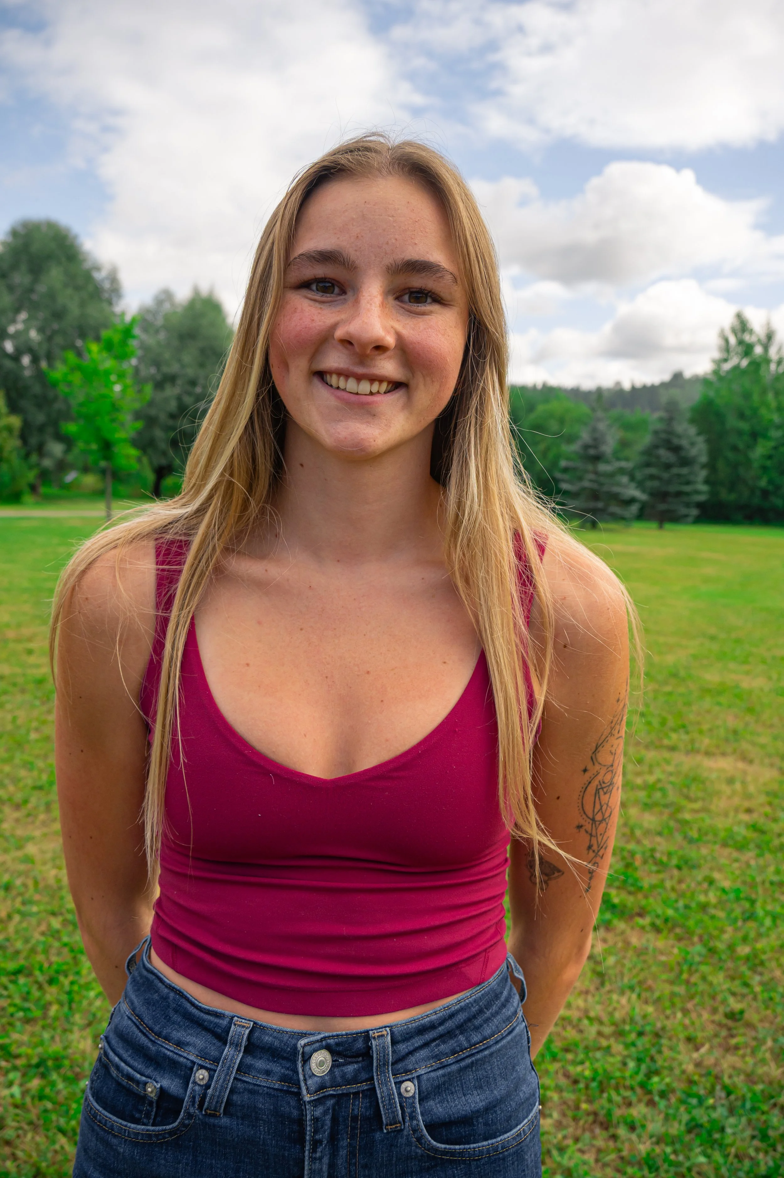 A young woman with long blonde hair smiling outdoors. She is wearing a sleeveless maroon top and denim shorts, and has a tattoo on her left arm. The background features green grass and trees under a partly cloudy sky.