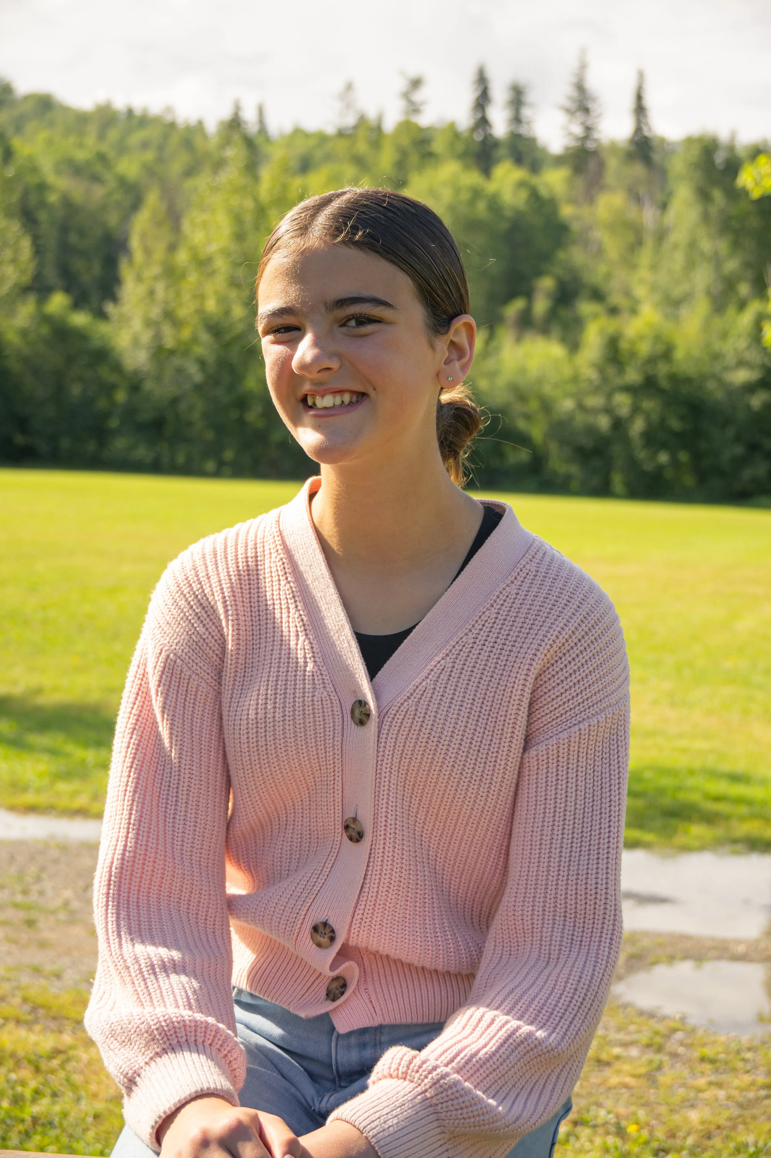 A young woman with dark hair tied back in a ponytail, wearing a pink cardigan with large buttons, smiling outdoors in a park with green trees and grass in the background.