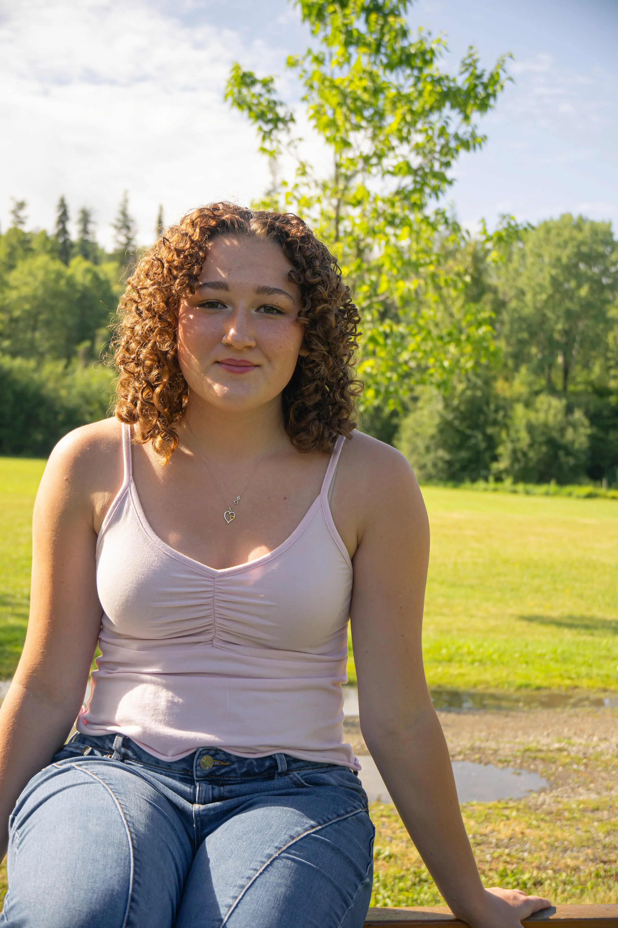 Young woman with curly brown hair sitting on a wooden bench outdoors on a sunny day, wearing a light pink tank top and blue jeans, surrounded by green trees and grass.