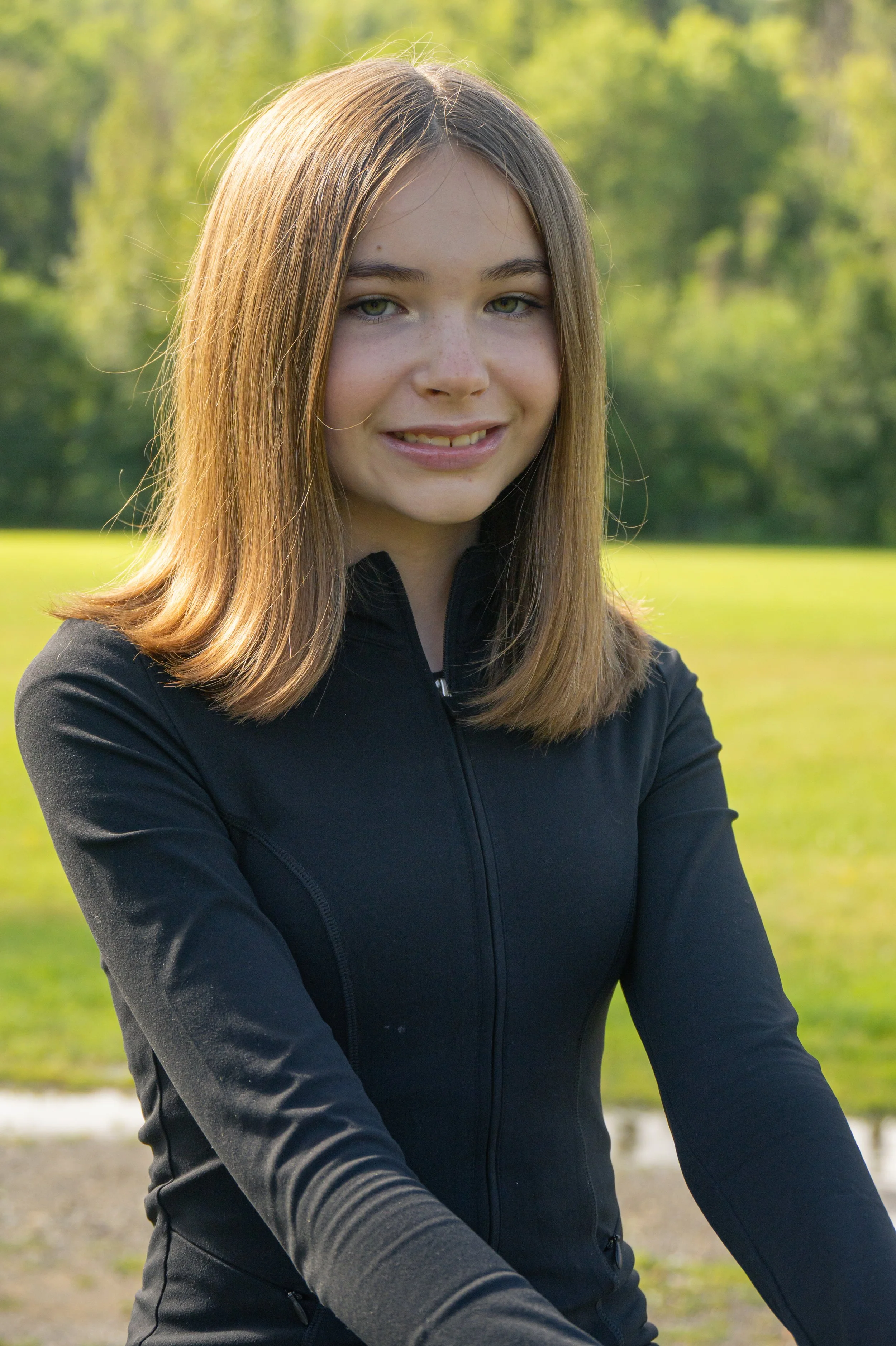 Young woman with light brown hair, green eyes, and freckles, smiling and wearing a black athletic jacket outdoors in a park.