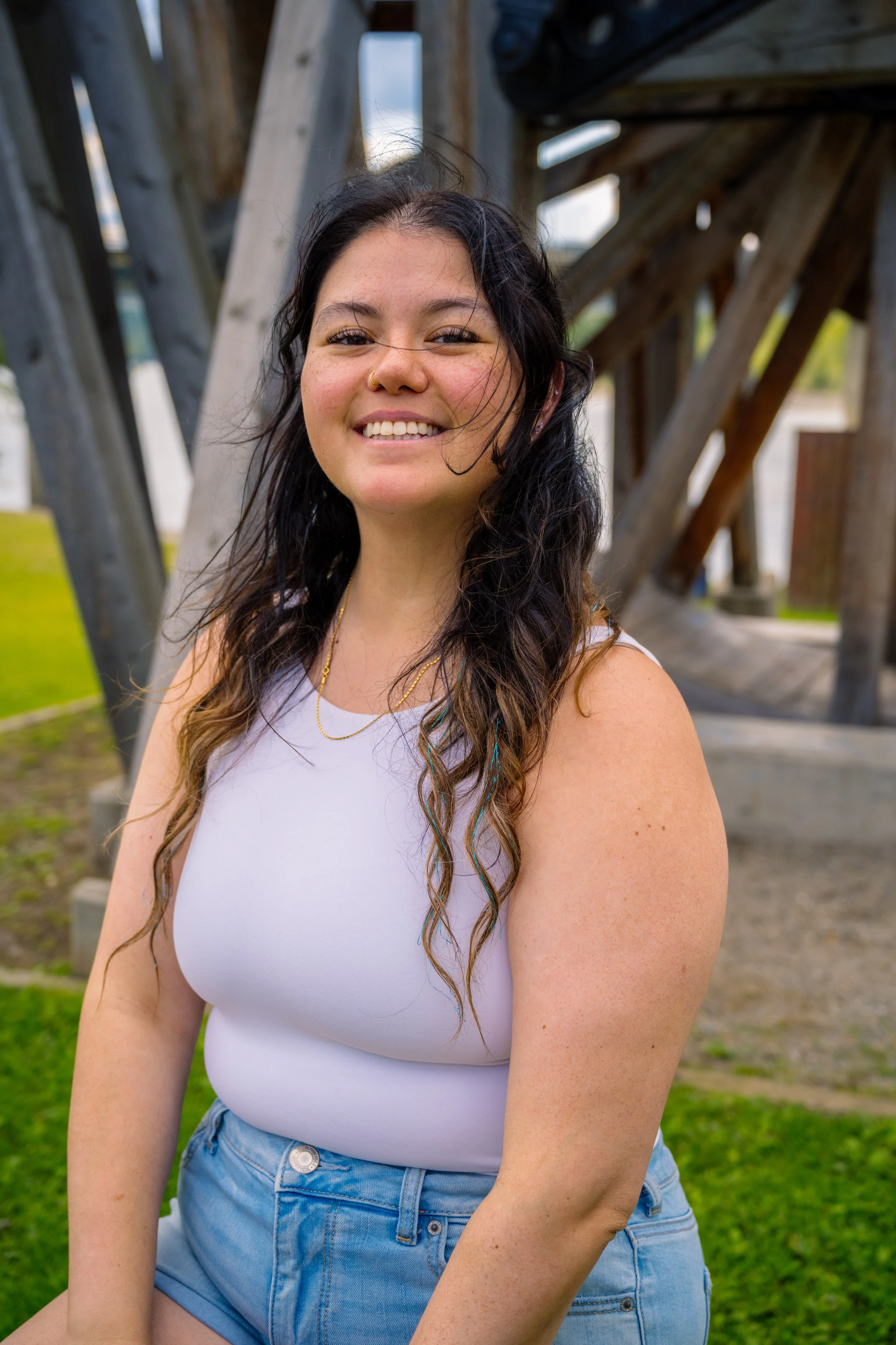 A young woman with long wavy dark hair standing outdoors in front of a wooden structure, smiling at the camera. She is wearing a white sleeveless top and light blue denim shorts.
