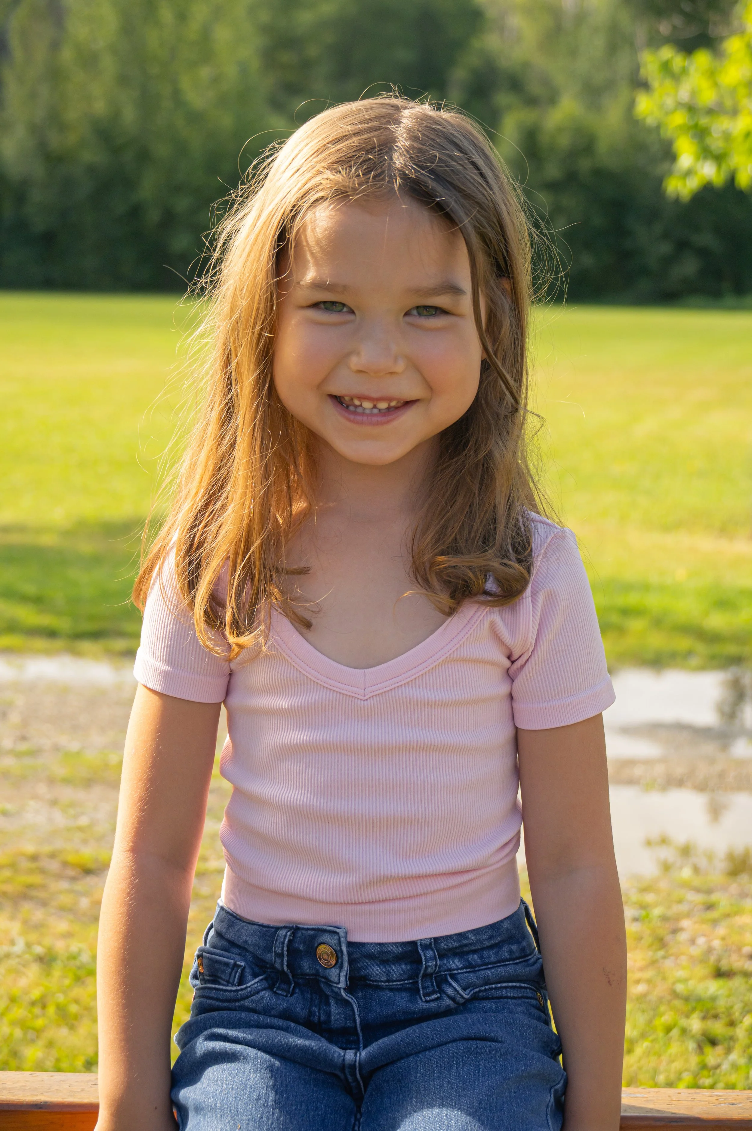A young girl with long light brown hair, wearing a light pink short-sleeve shirt and dark blue jeans, standing outdoors on a sunny day with green grass and trees in the background.