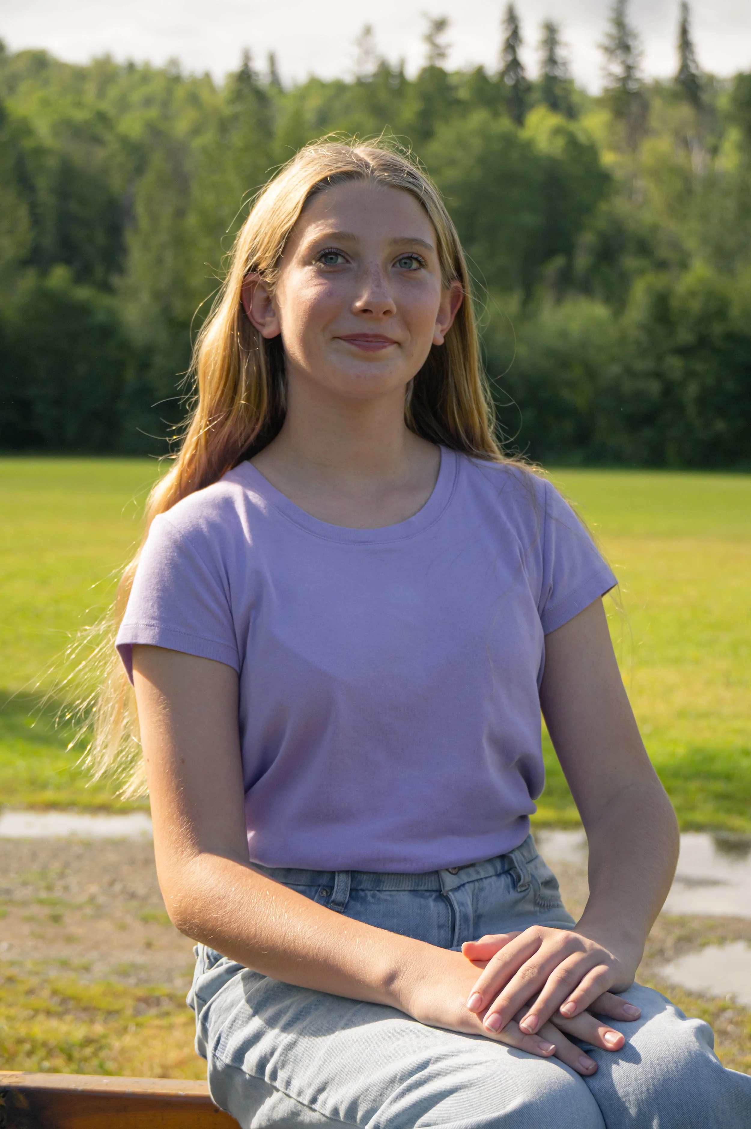 A young girl with long blonde hair and blue eyes sitting outdoors on a bench, wearing a lavender t-shirt and light-colored jeans, with a background of green trees and a grassy field.