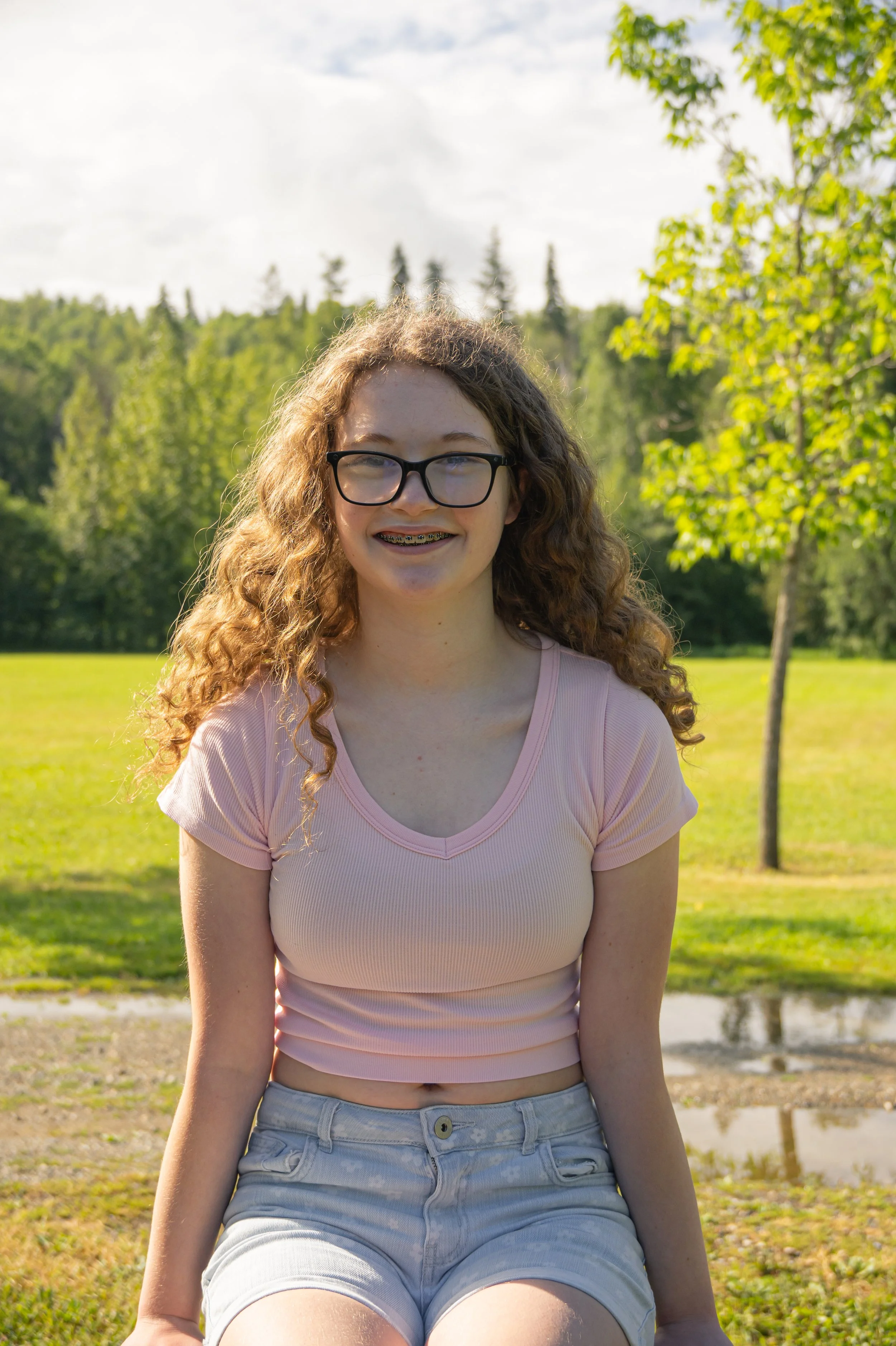 A young woman with curly red hair and glasses sitting outdoors in a park on a sunny day, smiling at the camera, wearing a pink T-shirt and light blue shorts, with trees and grass in the background.
