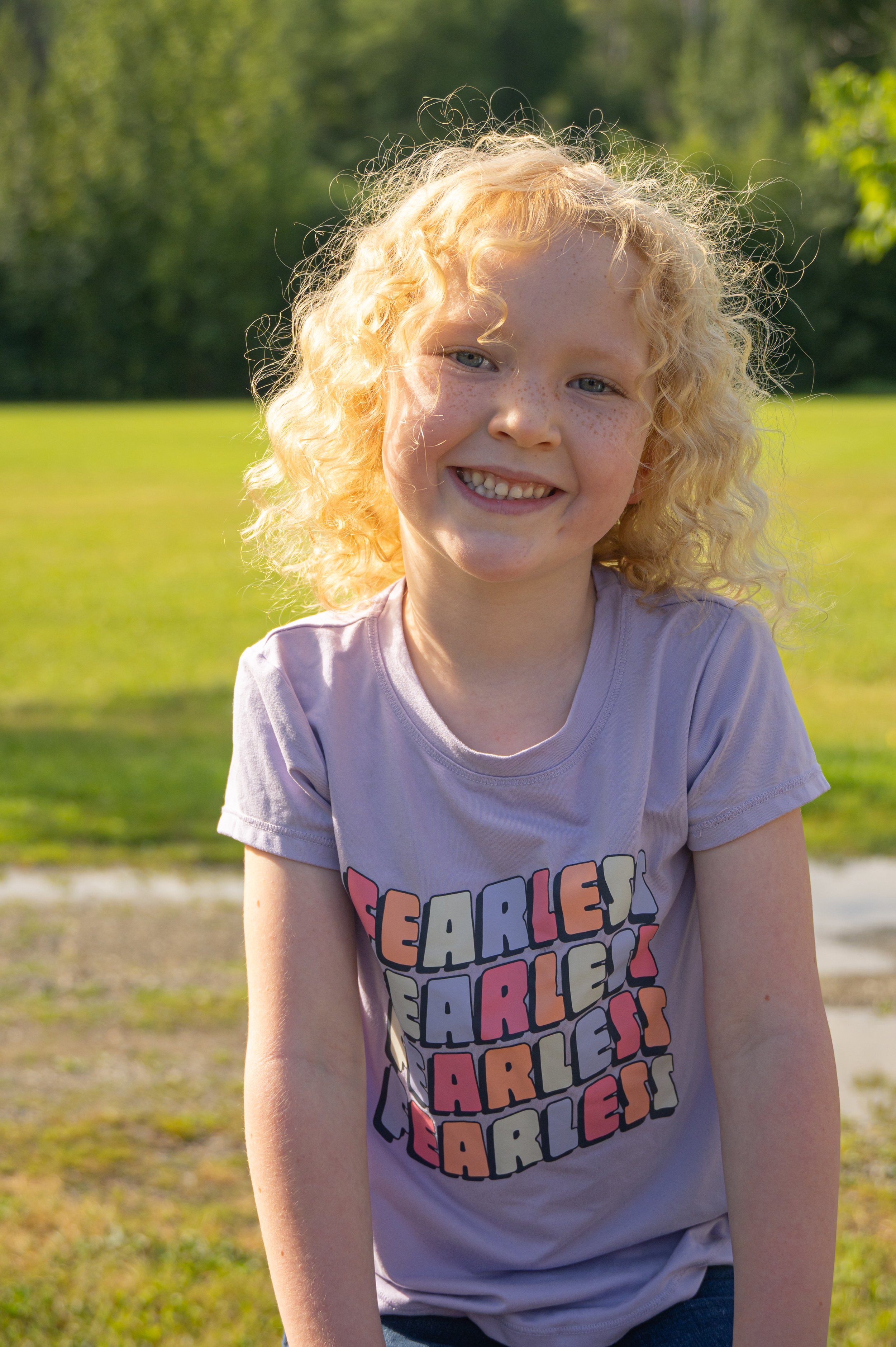 A smiling young girl with curly blonde hair and freckles, wearing a pink T-shirt with colorful text, standing outdoors in a sunny park with green trees and grass in the background.