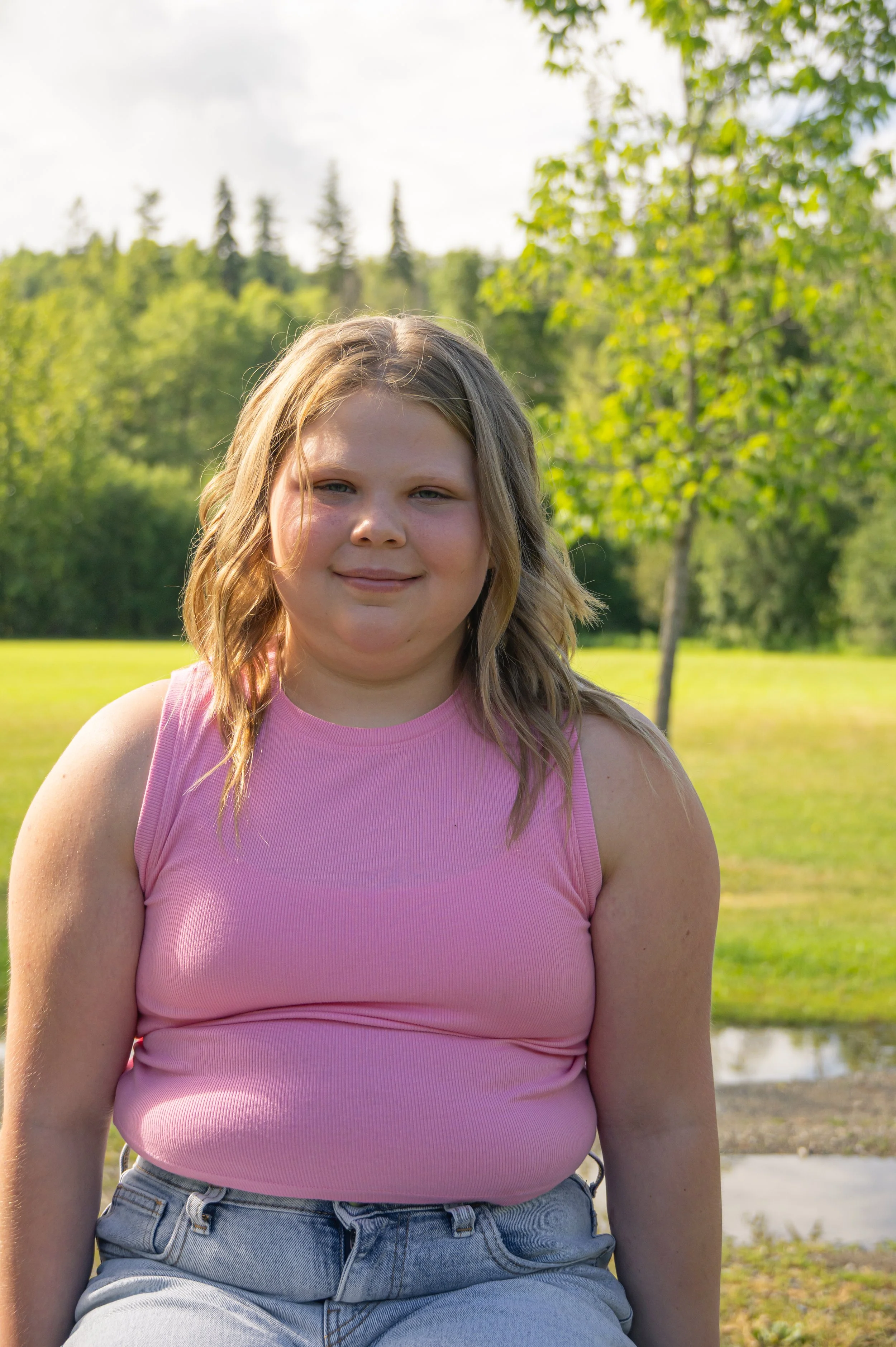 A young girl with blonde hair wearing a pink sleeveless top and jeans standing outdoors near greenery and trees.
