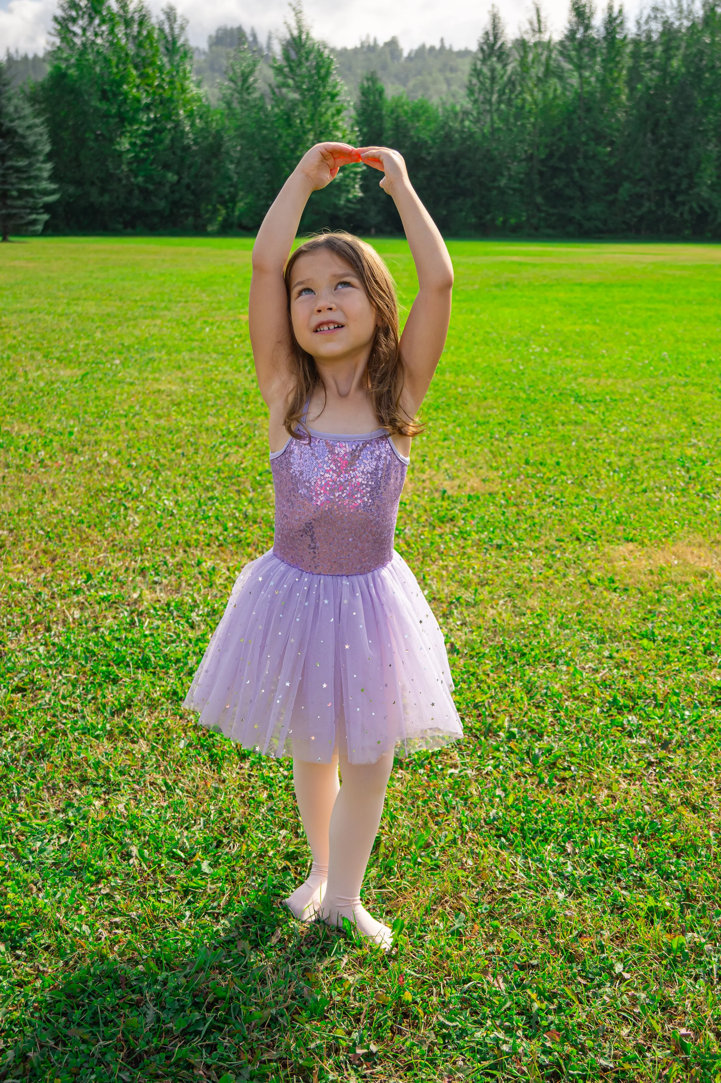 Young girl in a pink ballet tutu practicing a ballet pose outdoors on grass with trees in the background.