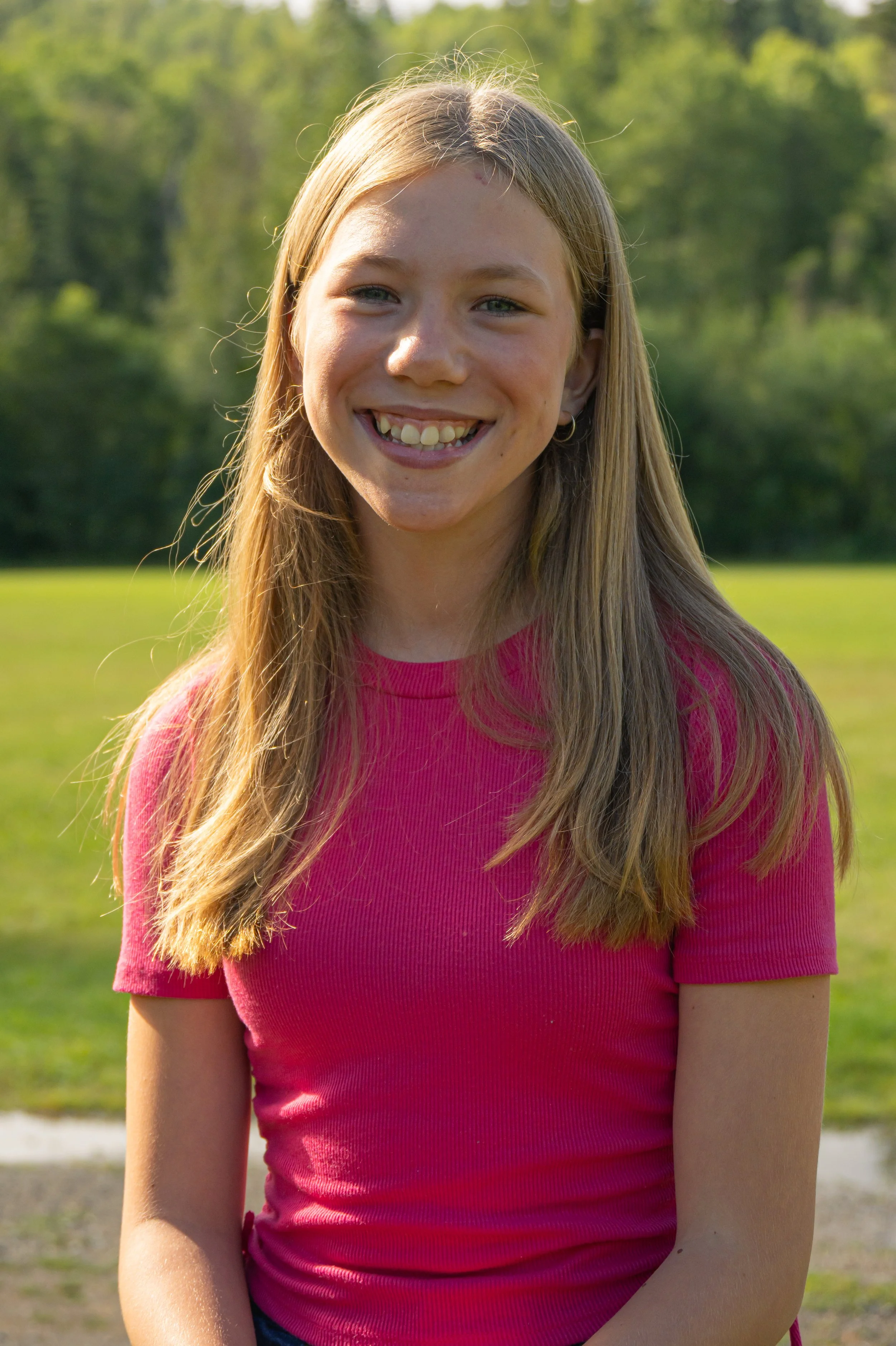 A young girl with long blonde hair, smiling outdoors in a park on a sunny day, wearing a pink shirt.