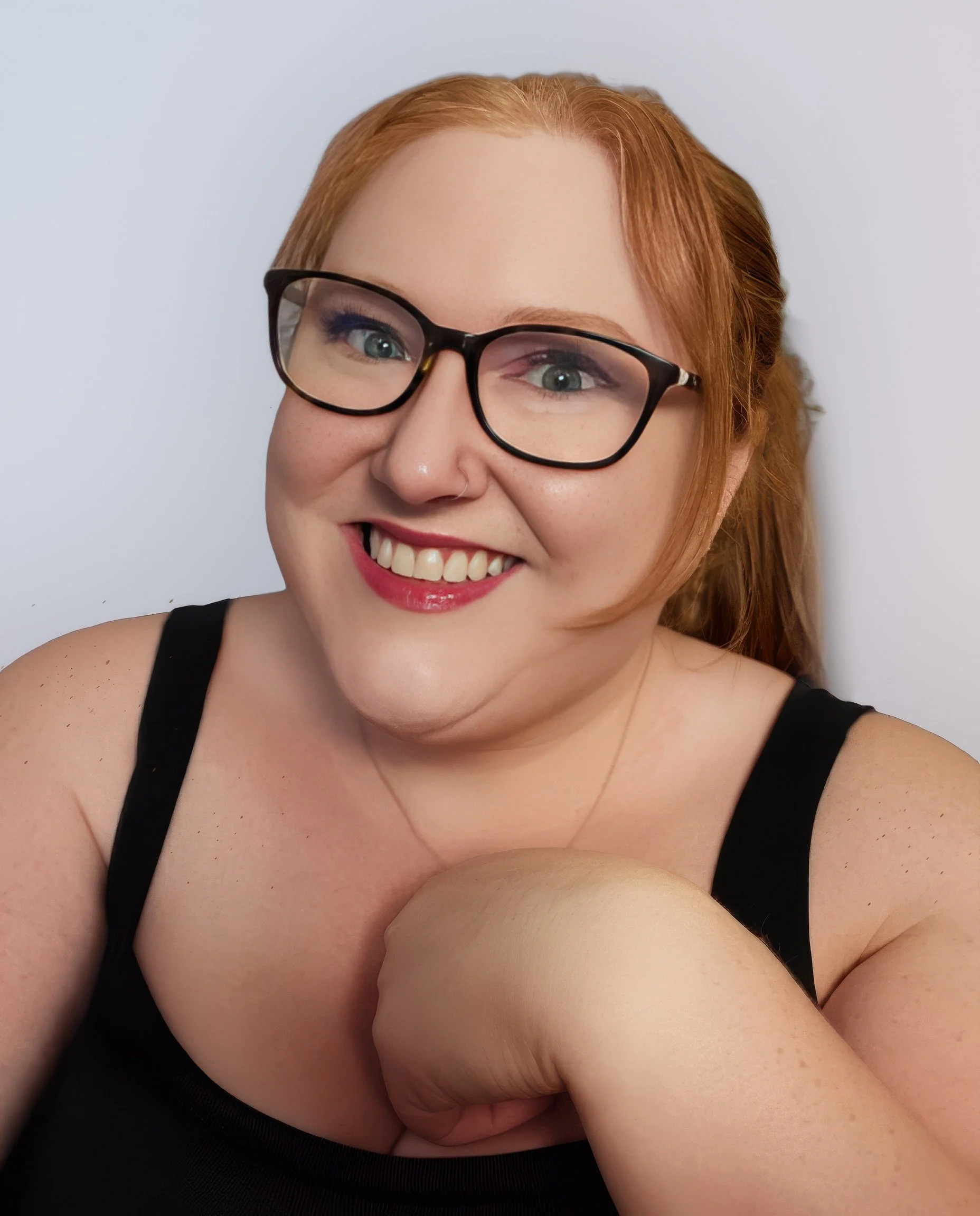 Close-up of a smiling woman with red hair, wearing glasses, in a black top, with her arms crossed, against a plain white background.