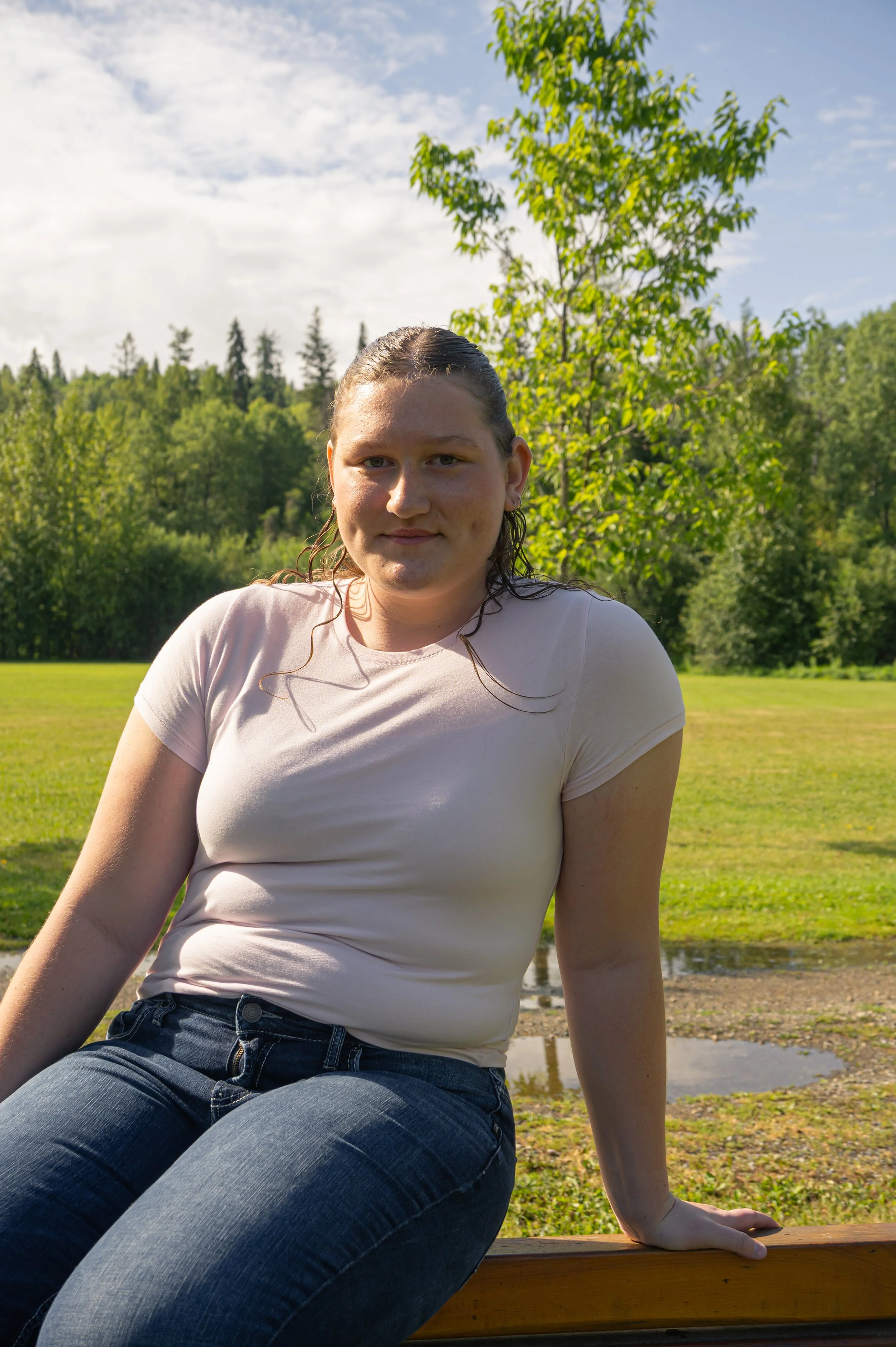 A woman with wet hair sitting on a park bench outdoors near a pond, with green trees and a partly cloudy sky in the background.