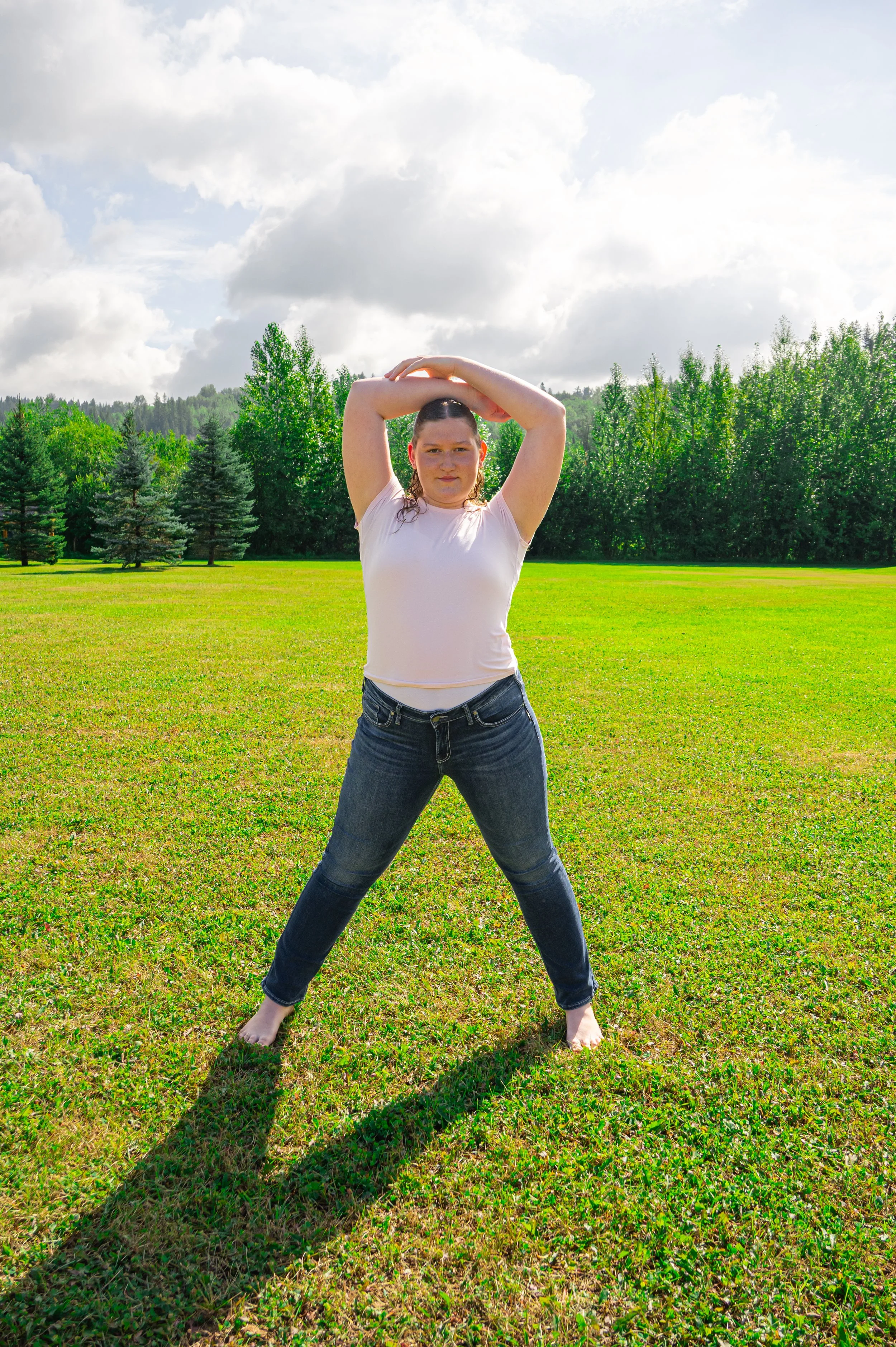 A young woman with wet hair and a white t-shirt stands barefoot on a grassy field with her arms raised above her head in a yoga pose, surrounded by green trees and a partly cloudy sky.