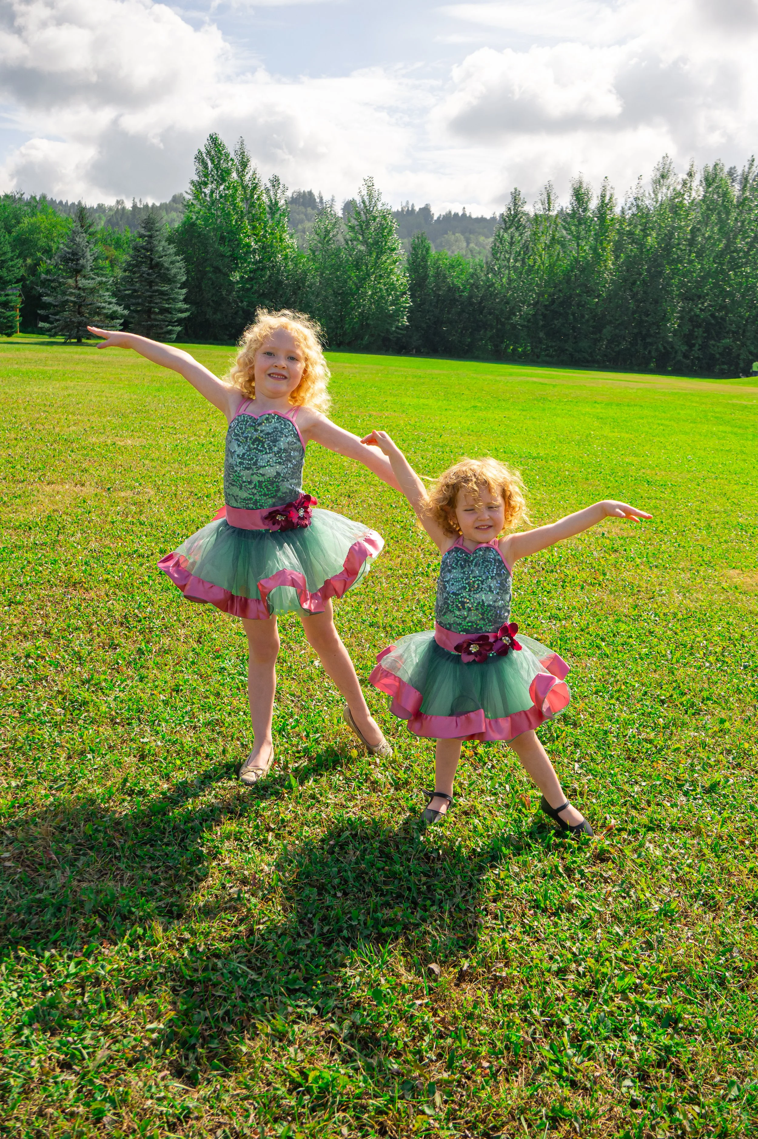 Two young girls in matching sparkly dance costumes with green tutus and pink accents pose on a grassy field under a partly cloudy sky, holding hands and smiling.