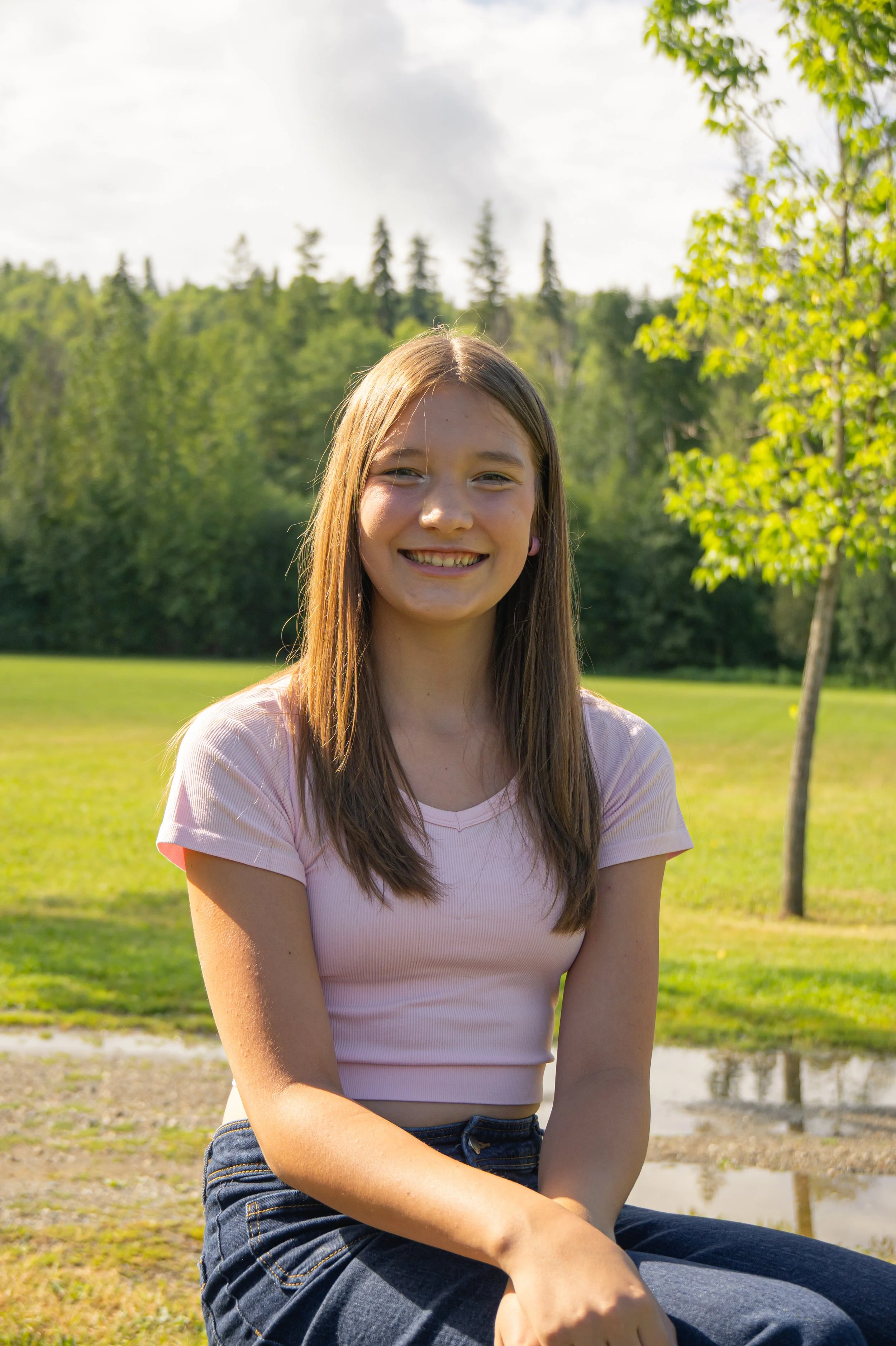 A young woman with long brown hair smiling outdoors in a park with green trees and a cloudy sky in the background.