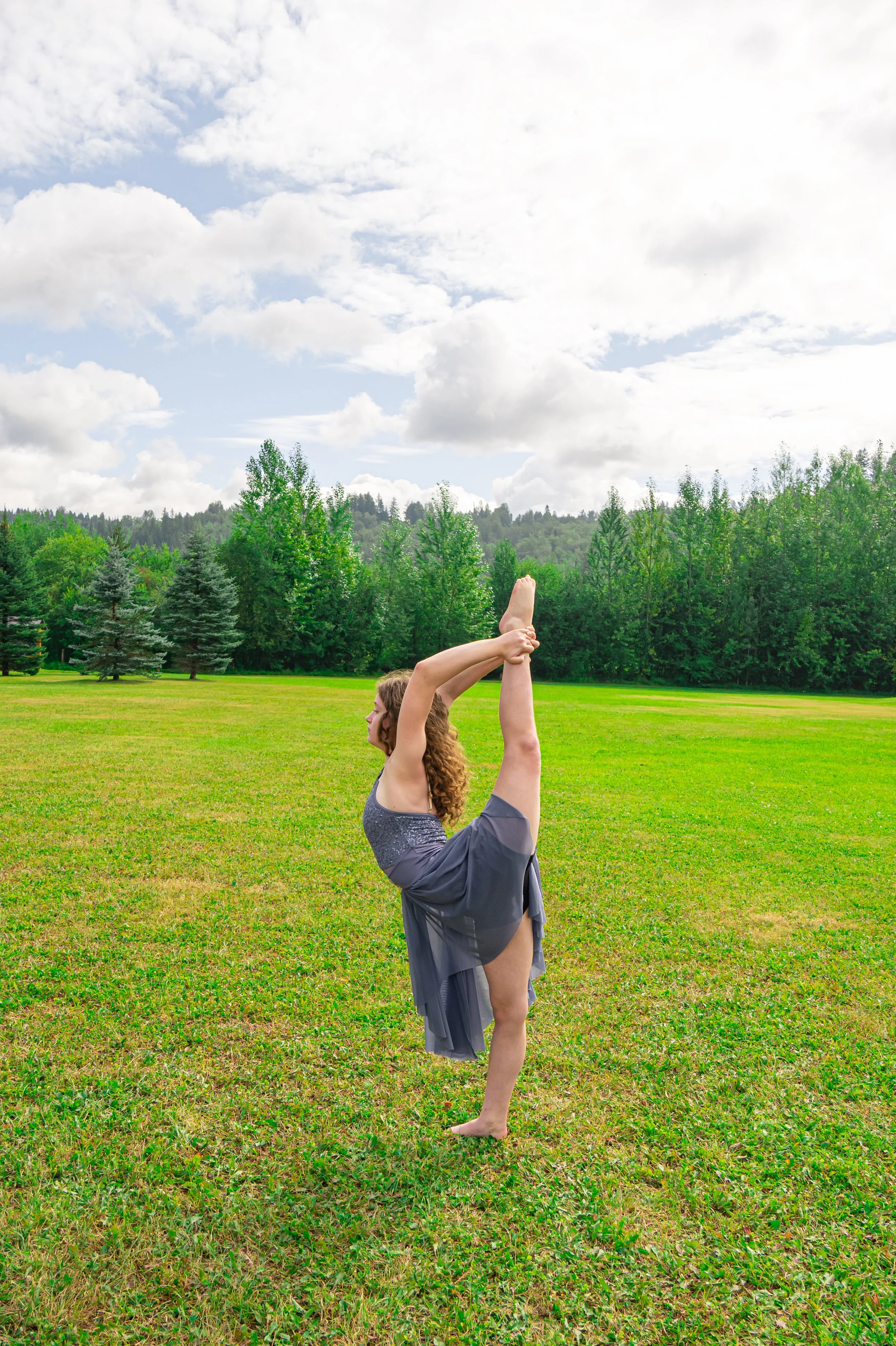A woman practicing yoga outdoors on a grassy field, performing a standing backbend pose with one leg lifted, arms reaching overhead, and trees in the background under a partly cloudy sky.