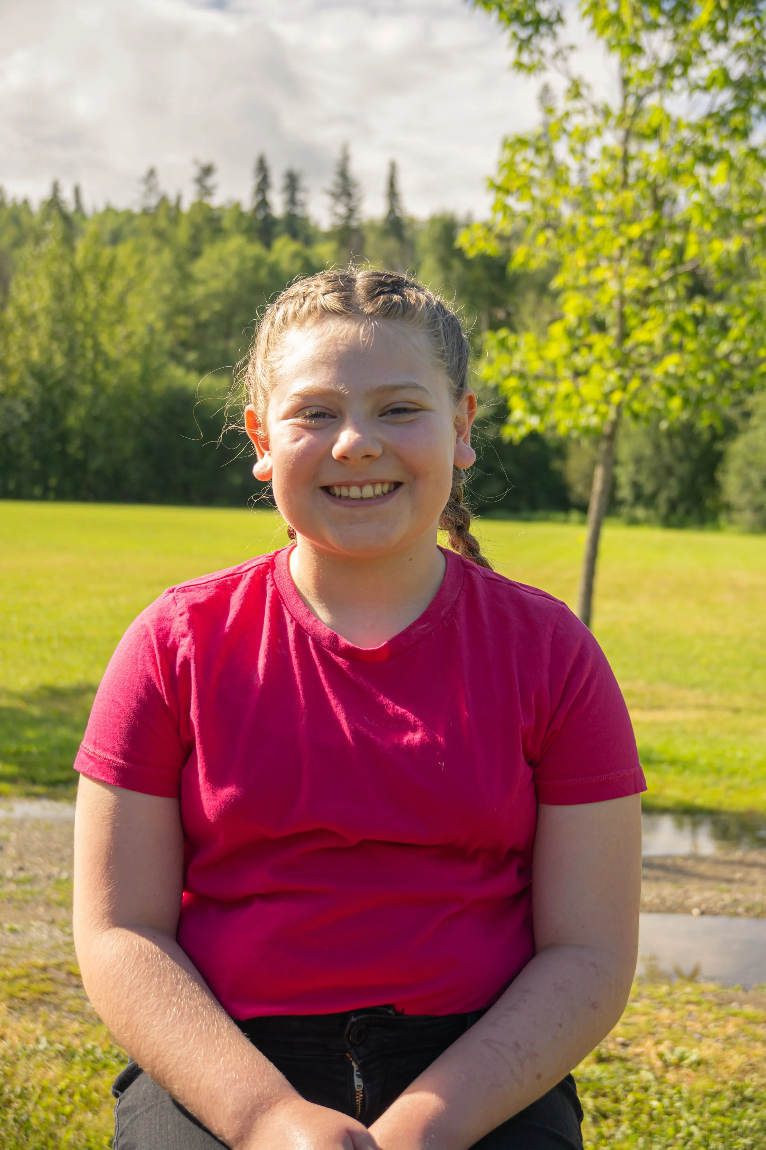 A young girl with braided hair wearing a red shirt sitting outdoors with a background of trees, grass, and cloudy sky, smiling at the camera.