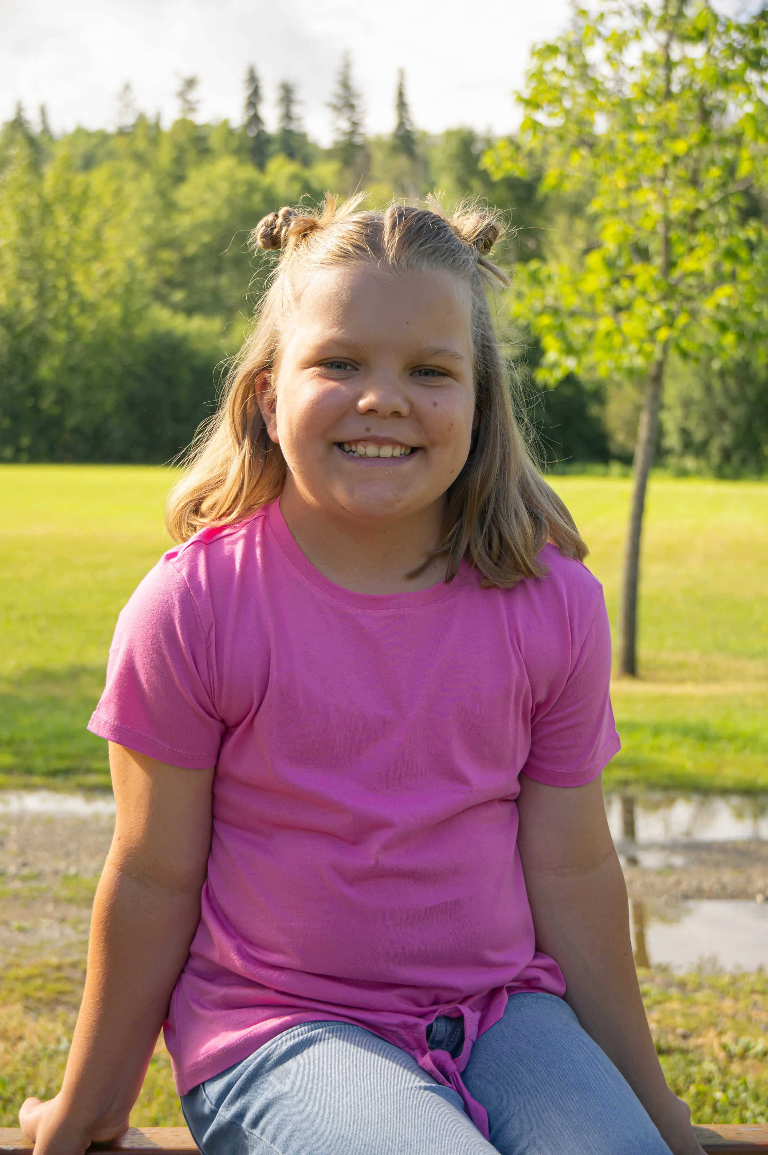 A young girl with blonde hair styled in two buns, smiling, sitting on a wooden surface outdoors with trees and green foliage in the background.