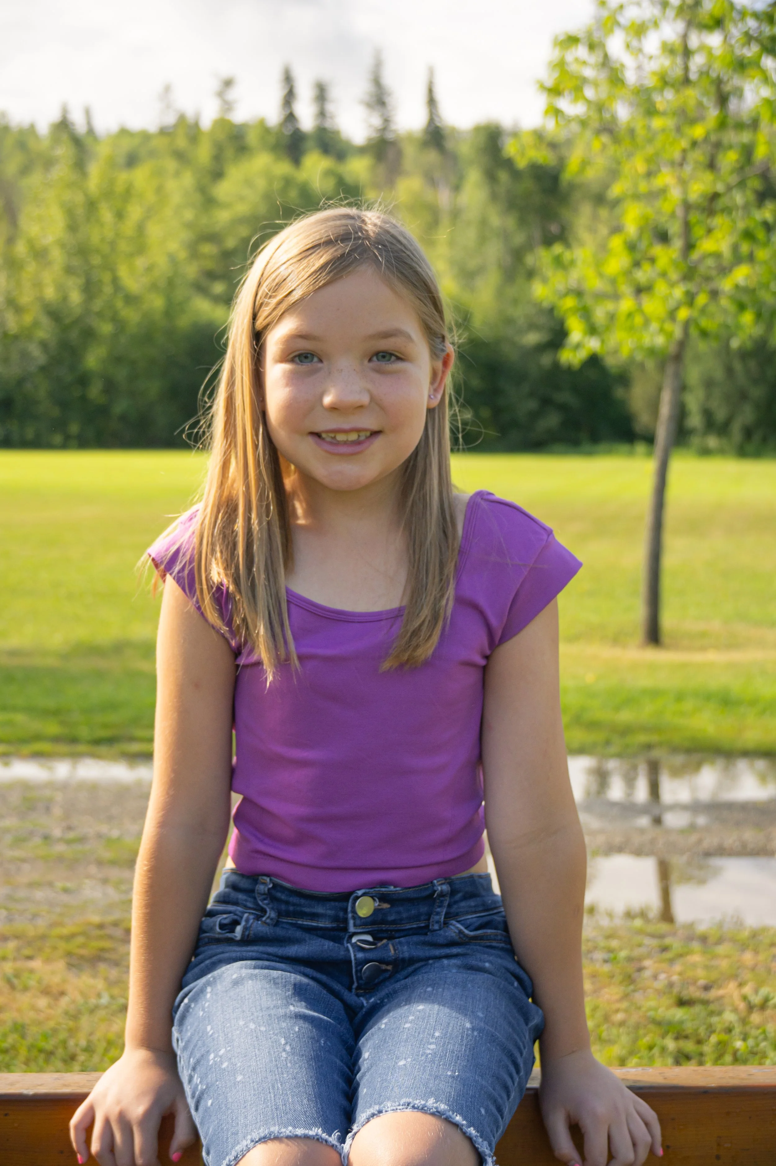 A young girl with long blonde hair, blue eyes, and freckles, wearing a purple t-shirt and denim shorts, sitting on a wooden bench outdoors with greenery and trees in the background.