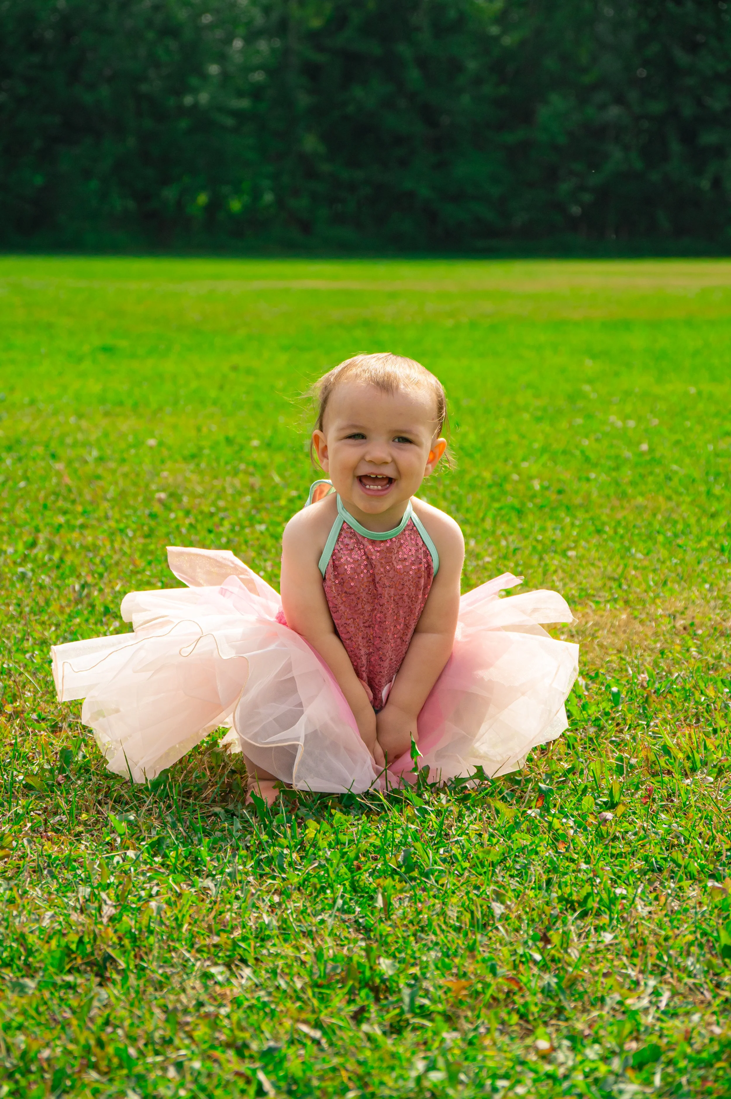 A young girl with a big smile, sitting on grass in a pink tutu dress with a glittery bodice, outdoors in a park or field, with a background of green trees.