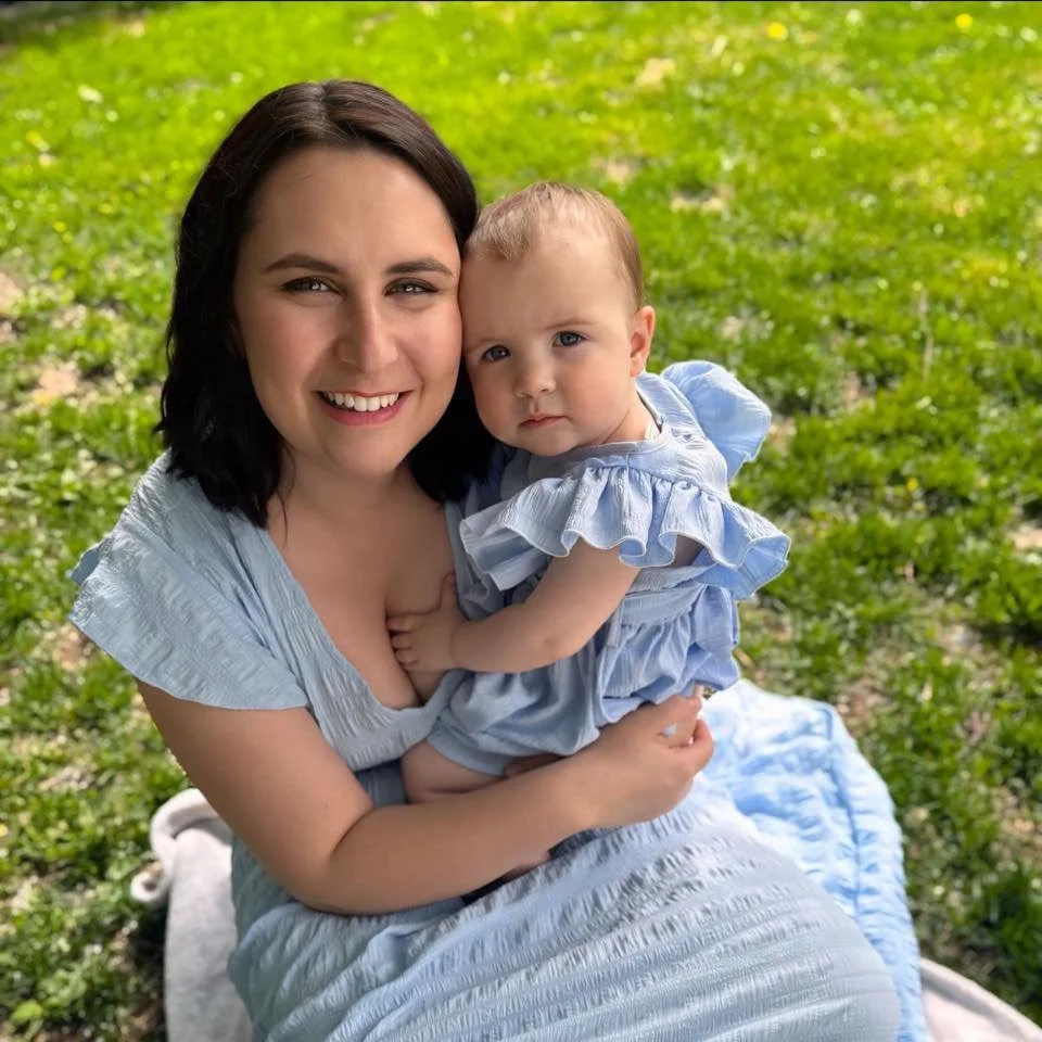 A woman with dark hair and light skin smiling while holding a young girl with light hair and light skin, both sitting outdoors on a blanket with green grass in the background.