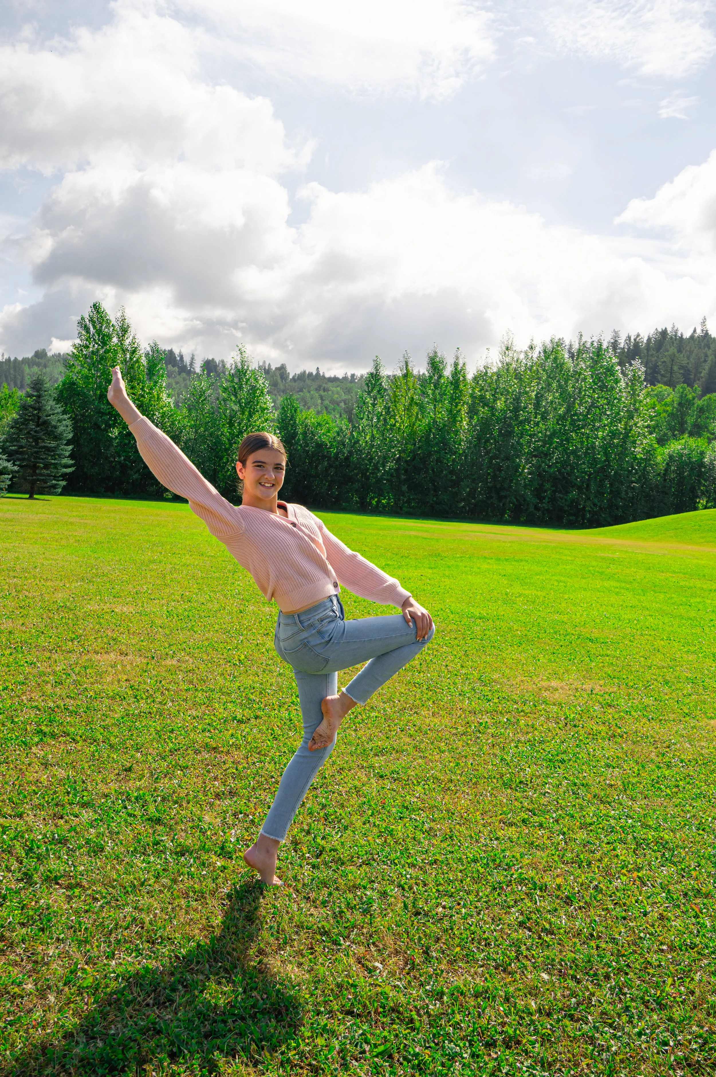 A young woman practicing yoga outdoors on a grassy field, balancing on one leg with her other leg bent and foot resting on her standing thigh, in a tree pose. She is smiling and wearing a light pink sweater and light blue jeans, with a backdrop of green trees and a partly cloudy sky.
