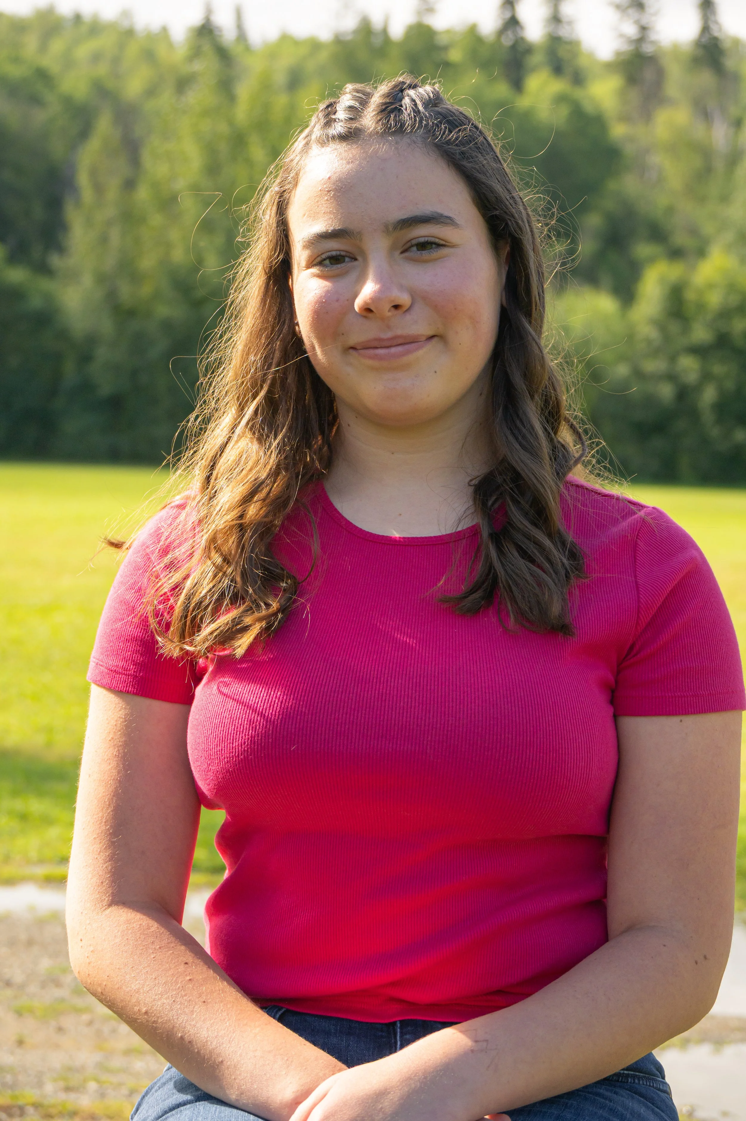 Close-up of a young woman with long, wavy brown hair, wearing a pink t-shirt, sitting outdoors on a sunny day with green trees in the background.