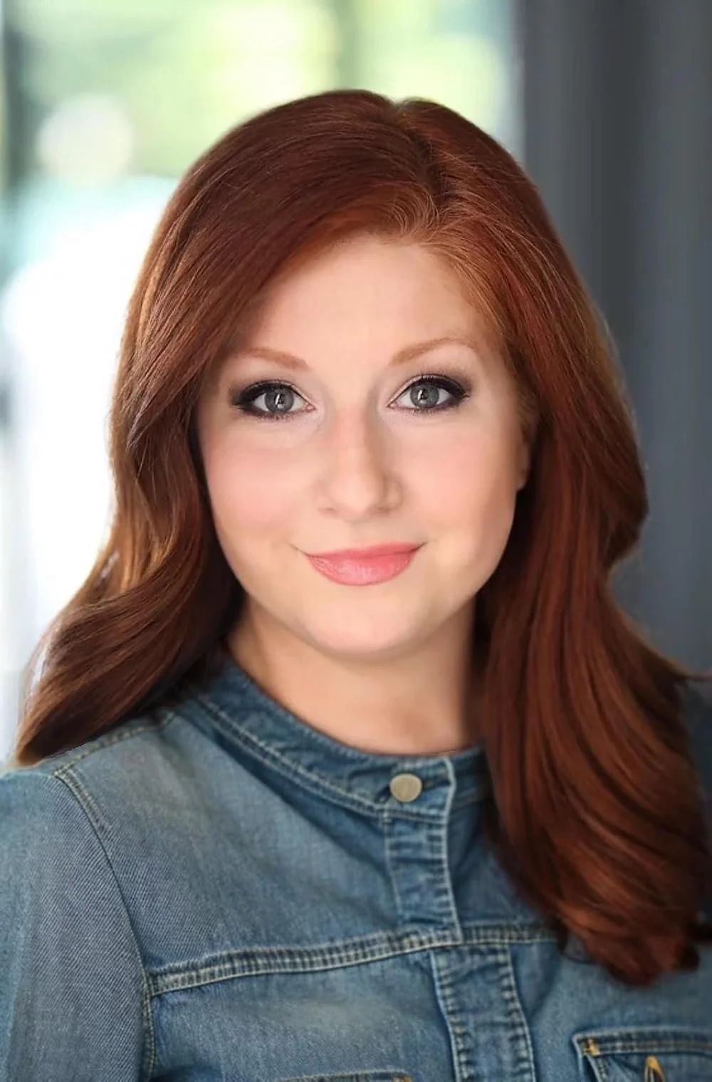 Close-up portrait of a young woman with red hair, blue eyes, and fair skin, wearing a denim jacket, smiling gently.