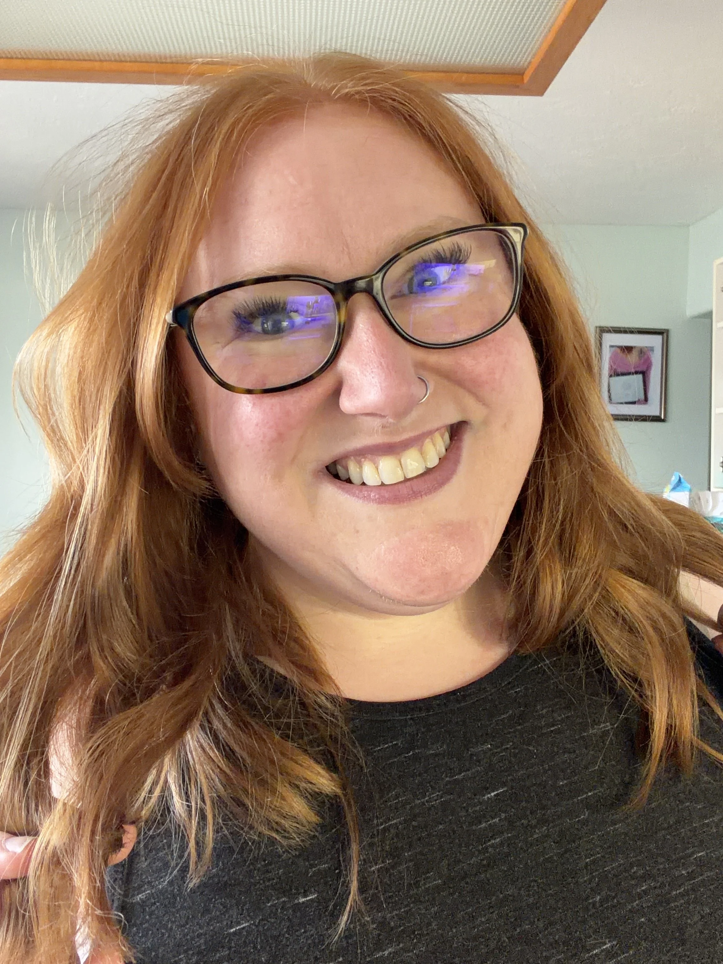 A woman with long red hair, wearing black-rimmed glasses, smiling, and wearing a black top. The background features a framed picture on a green wall.