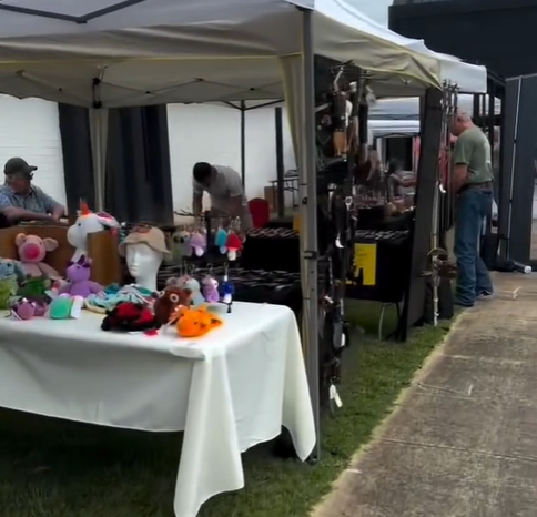 A street market stall with plush toys and accessories for sale, set up under a white canopy with vendors and customers in the background.